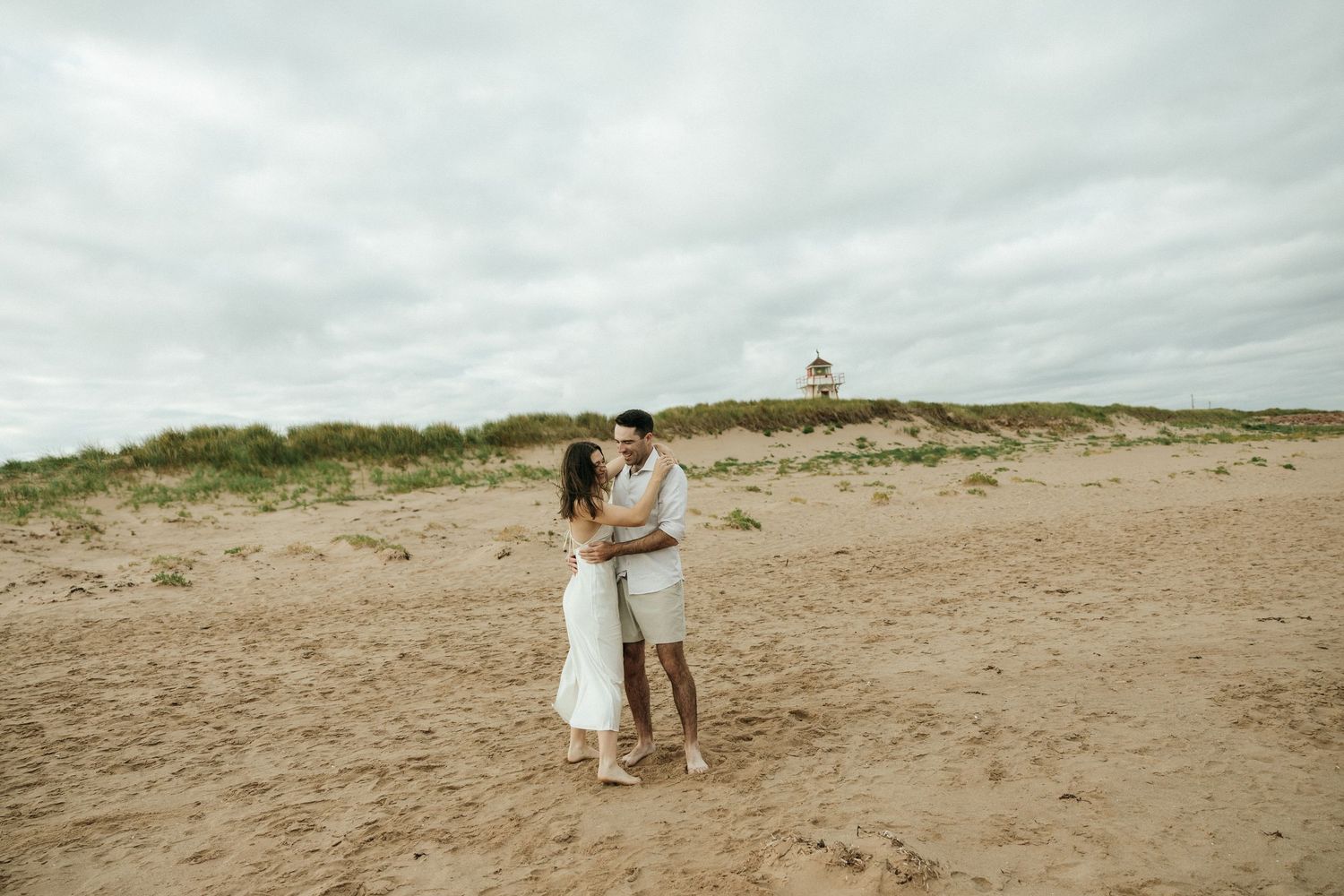 A couple stands together on a sandy beach with dunes and overcast skies in the background.