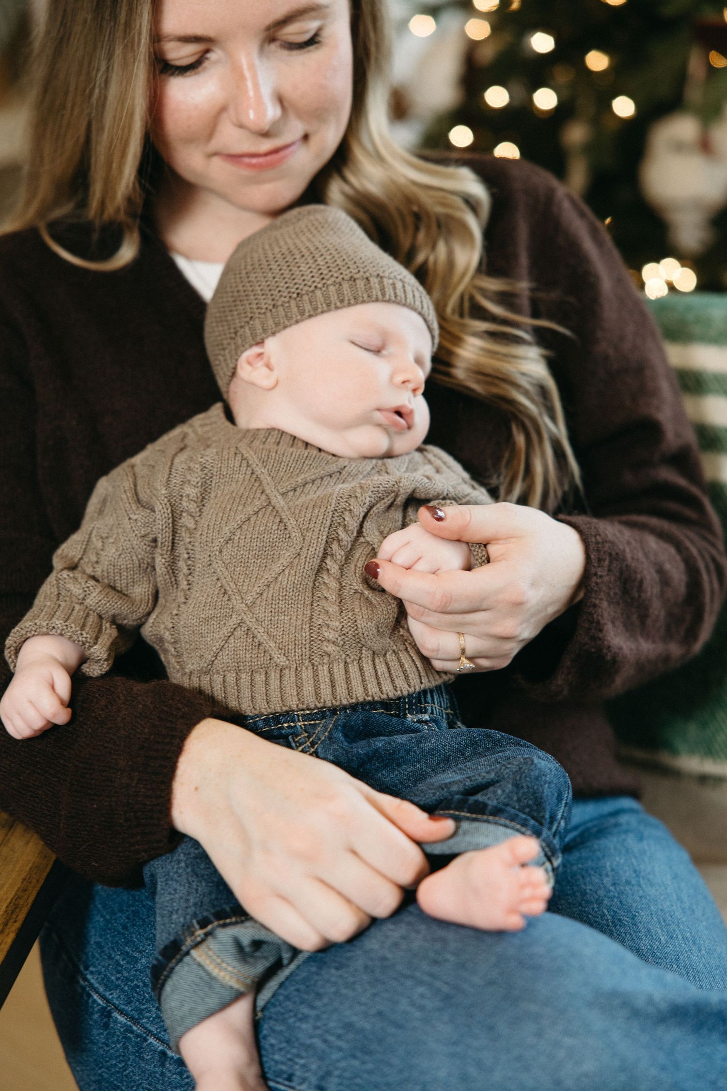 Baby wearing brown knit sweater and jeans held in loving arms against dark clothing.