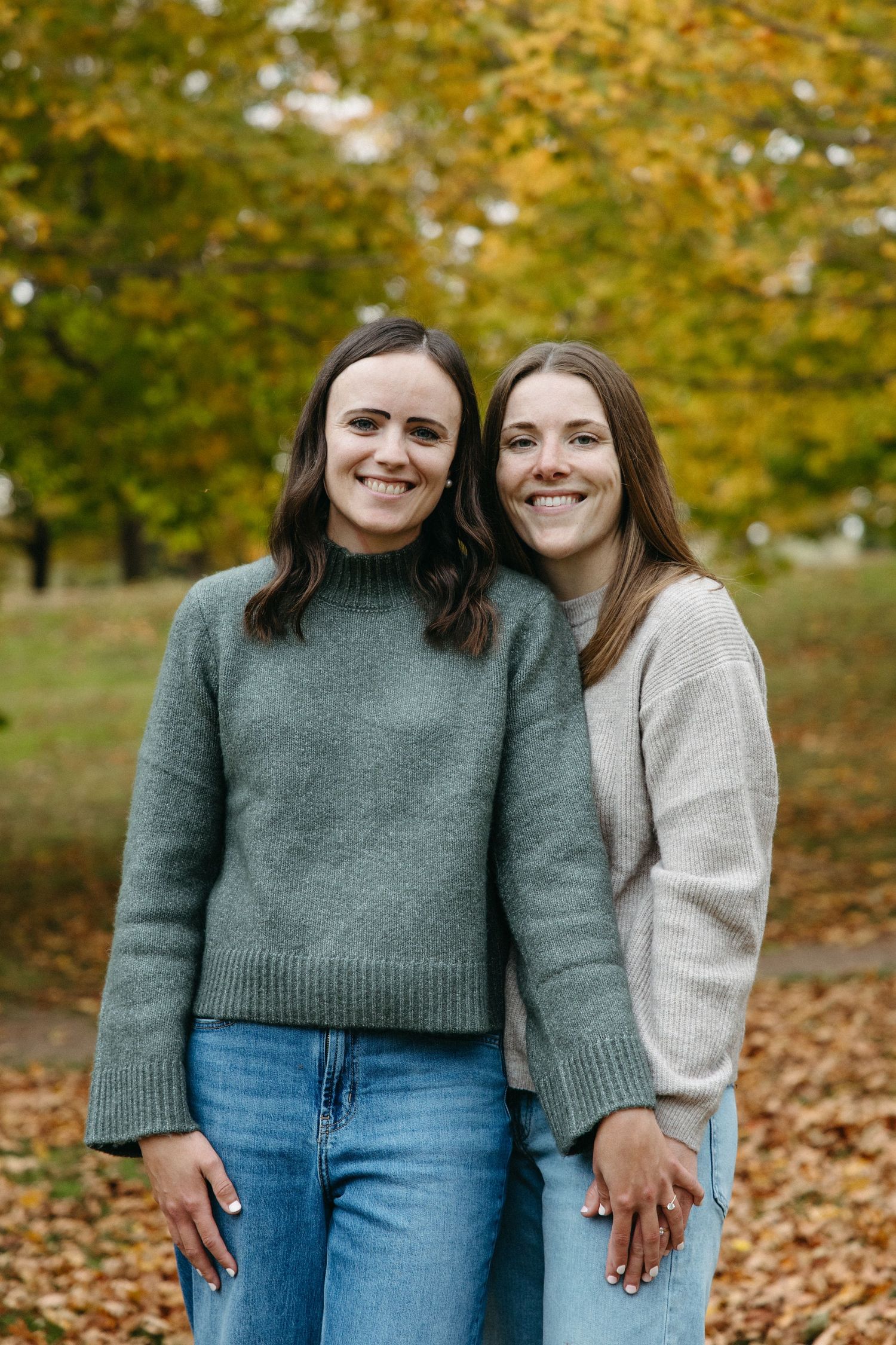 Two smiling women in cozy sweaters and jeans pose close together outdoors among golden autumn trees and fallen leaves.