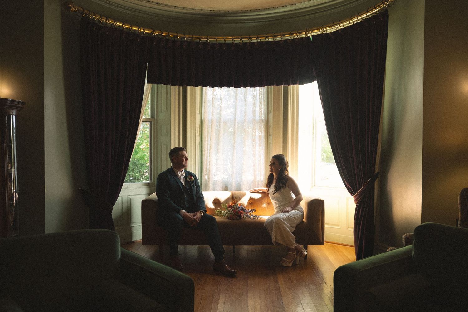 Bride and groom sit together on a vintage sofa with a bouquet in an elegant room with dark drapes and bay windows.