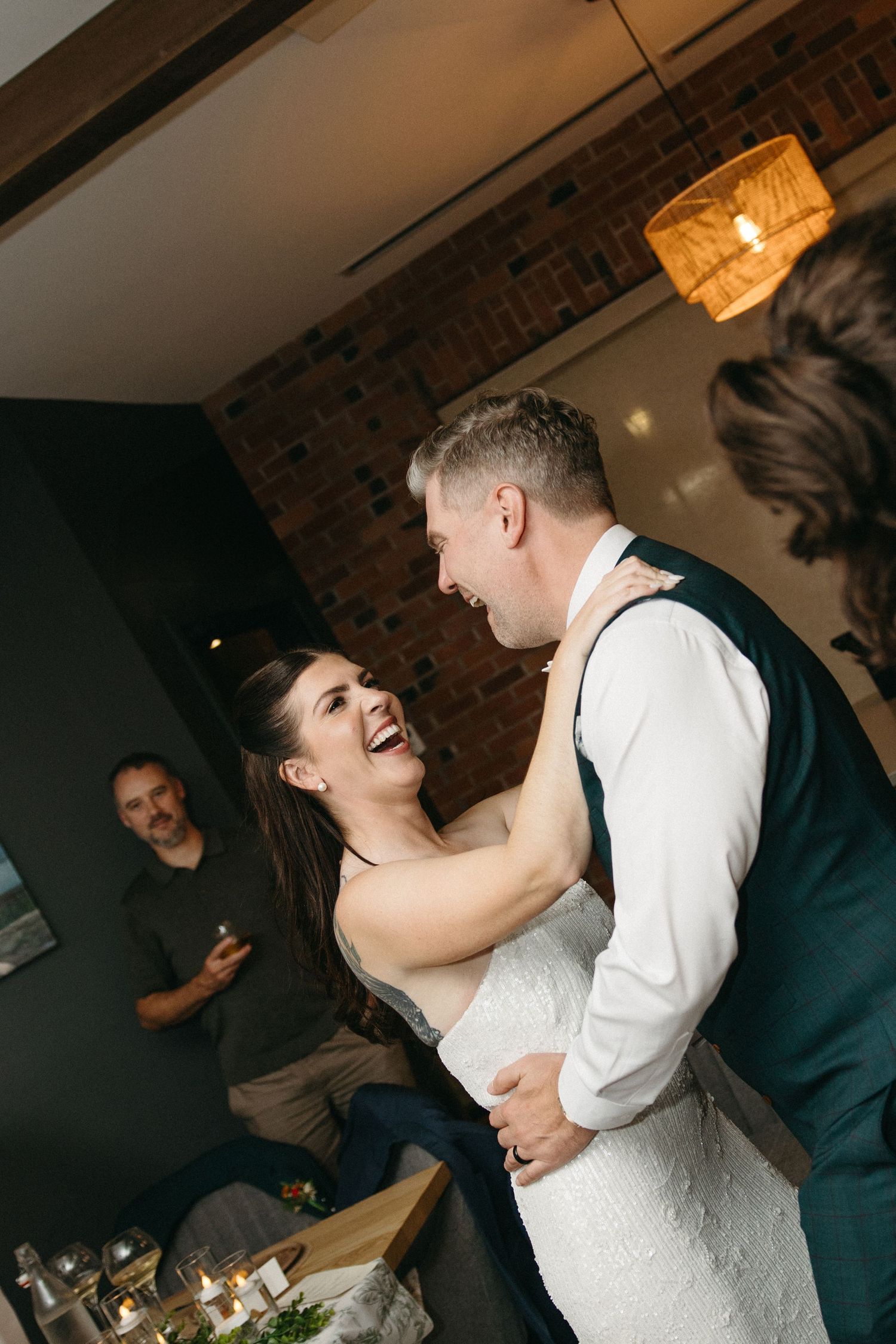 Smiling bride in a lace gown dances with her groom in a vest at an indoor wedding reception with brick walls and a guest watching.
