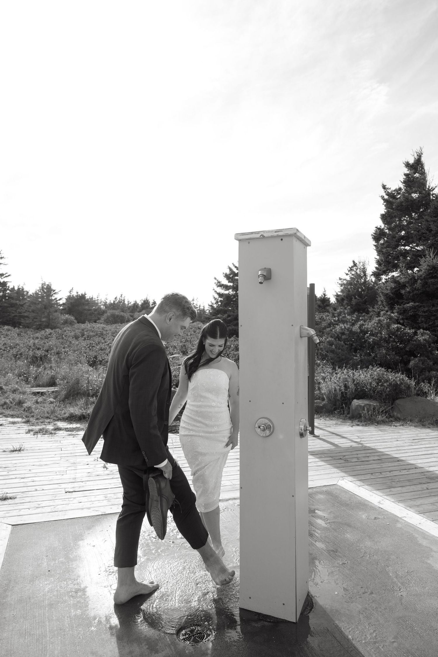 Black and white photo of a barefoot couple in formal attire rinsing their feet at an outdoor beach shower on a wooden boardwalk.