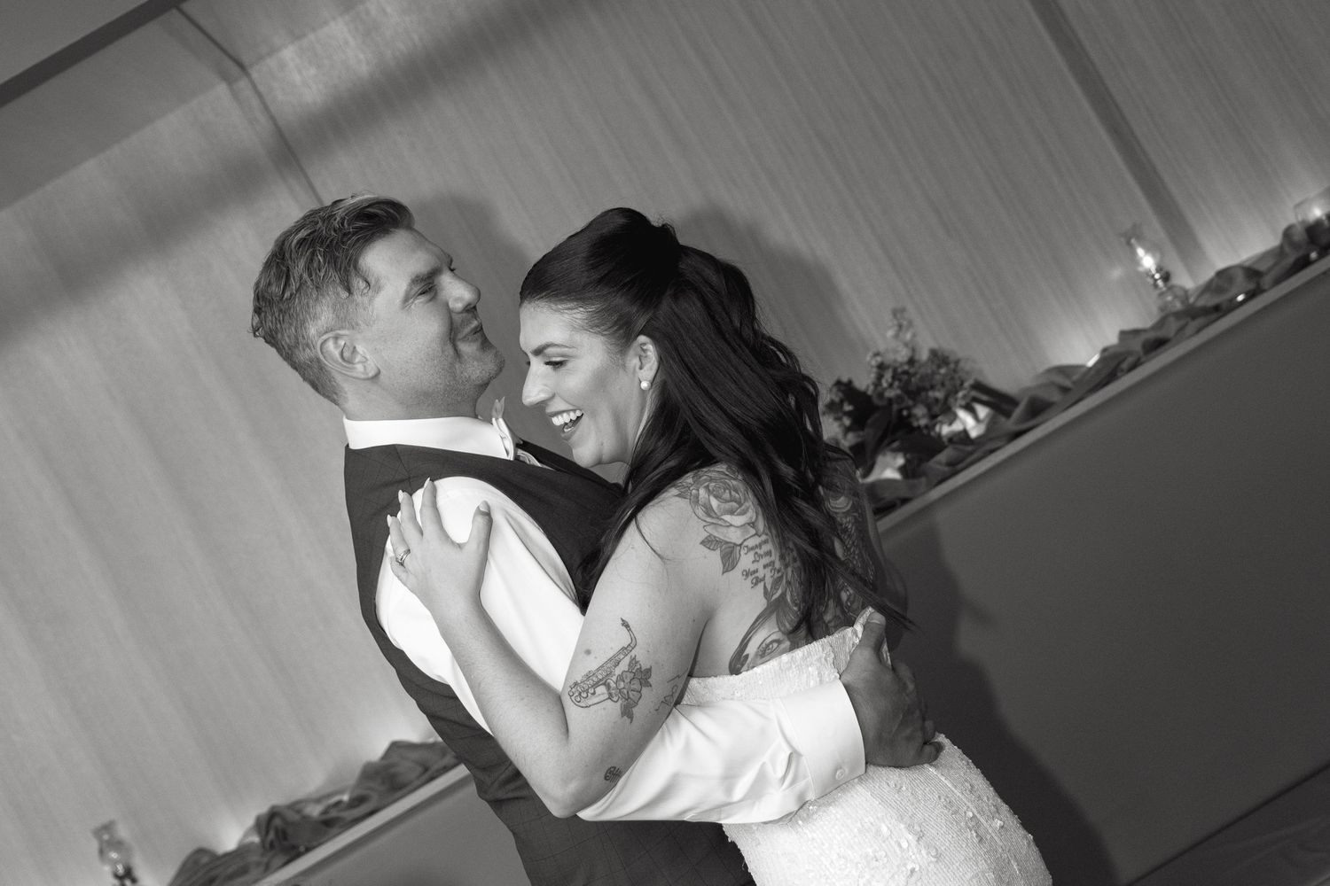 Black and white photo of a smiling bride with tattooed arms sharing a first dance with her groom at a wedding reception.