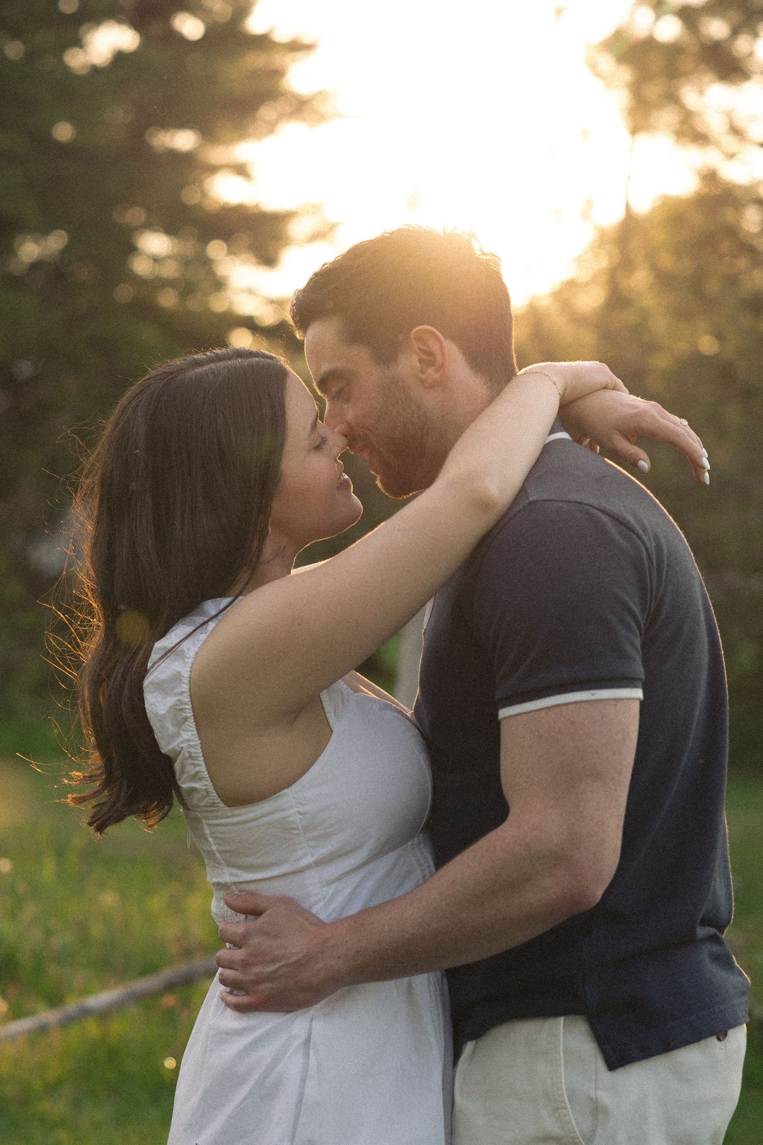 Couple sharing a romantic kiss outdoors at golden hour with warm sunlight glowing through trees behind them.