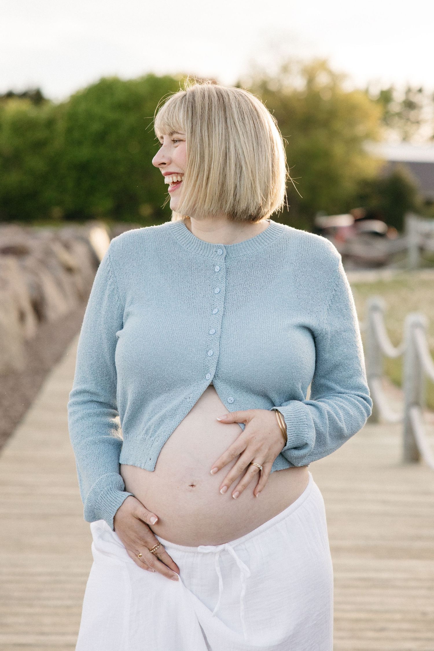 Smiling pregnant woman in blue cardigan and white skirt holds her bare baby bump outdoors on a wooden dock.