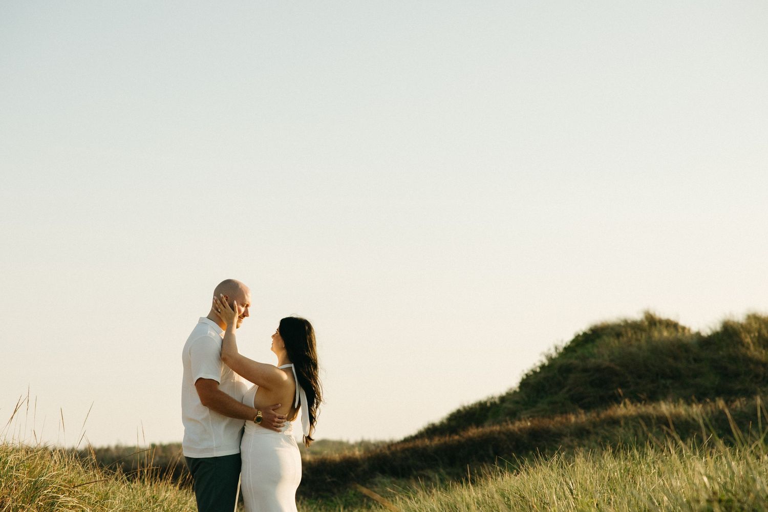 Couple sharing a tender moment on a grassy dune at golden hour.