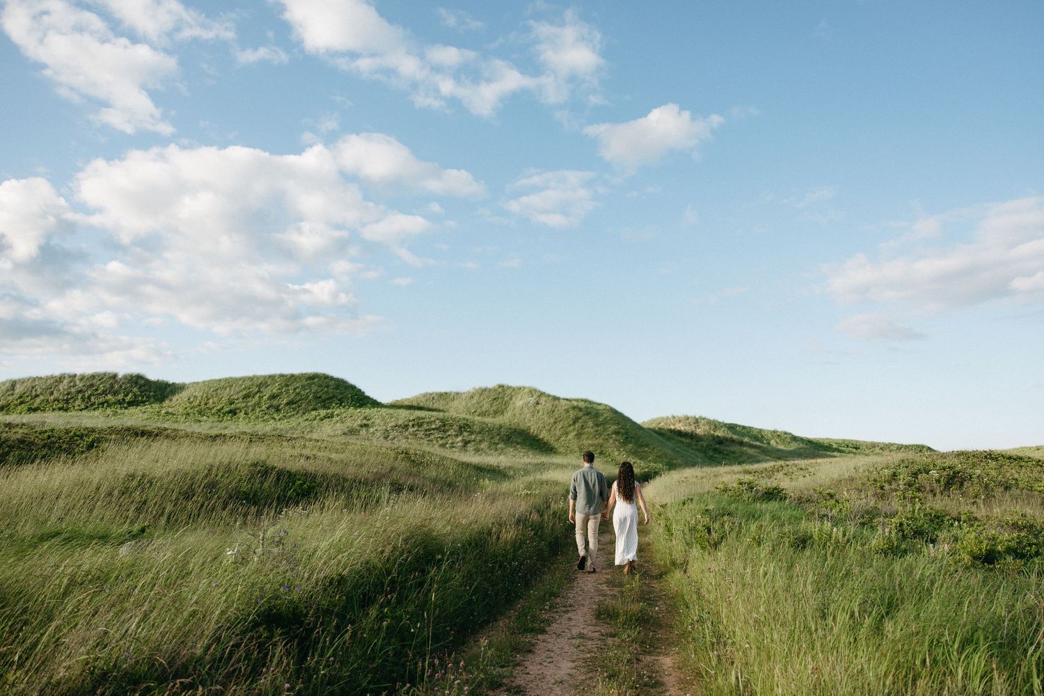 Two people walking along a winding dirt path through lush green rolling hills under a blue cloudy sky.