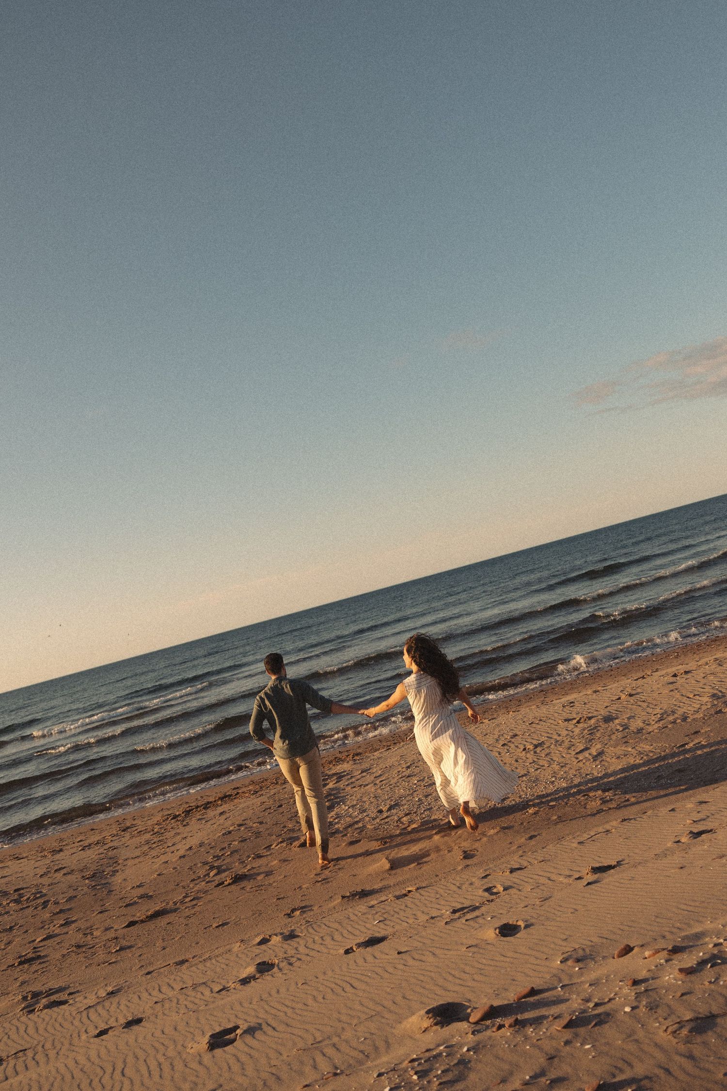 Couple holding hands walking toward ocean at sunset on sandy beach, woman in white dress and man in casual attire.