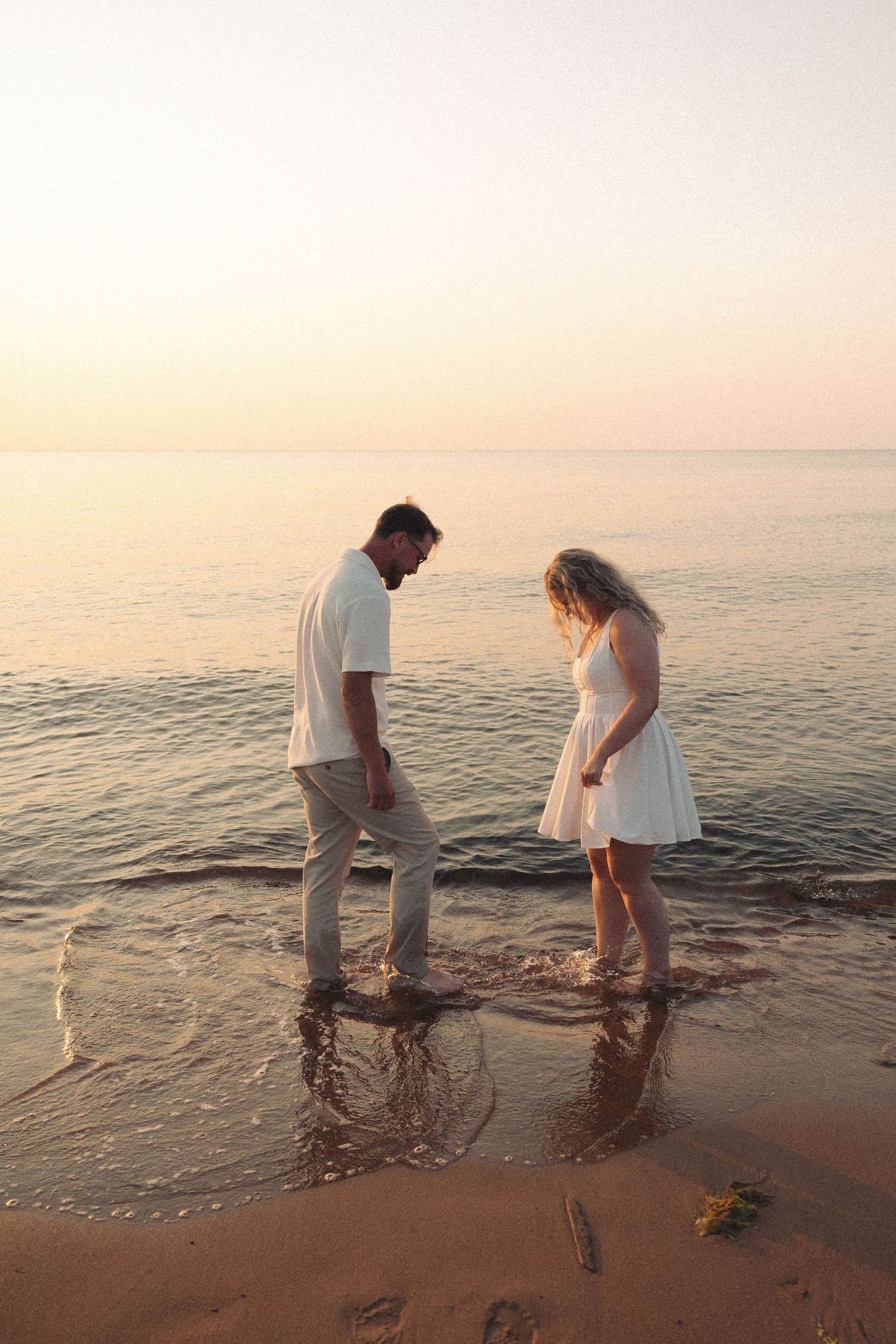 Couple standing barefoot in shallow ocean waves at sunset, both wearing white outfits on a sandy beach.