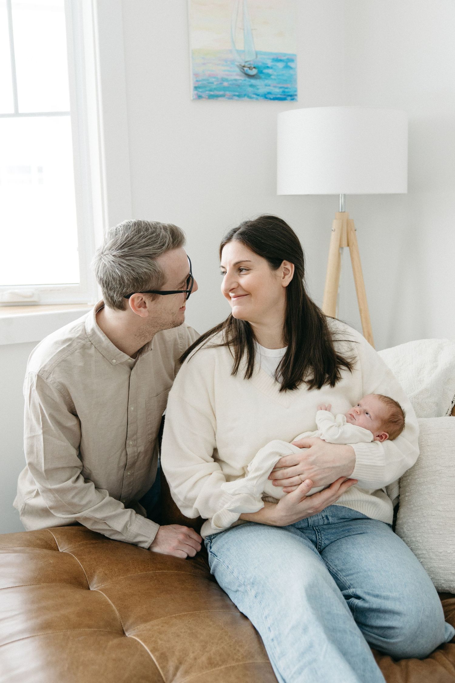 A couple in light colored sweaters shares tender moments with their newborn baby on a brown leather couch.