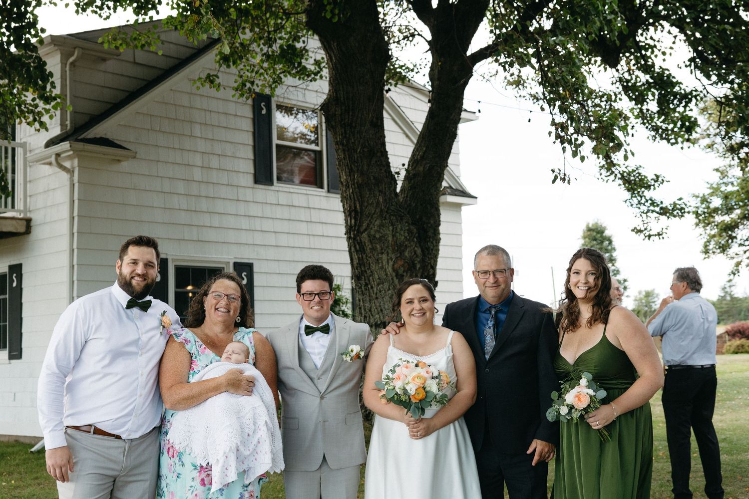Wedding party group photo with bride, groom, and family outside a white farmhouse.
