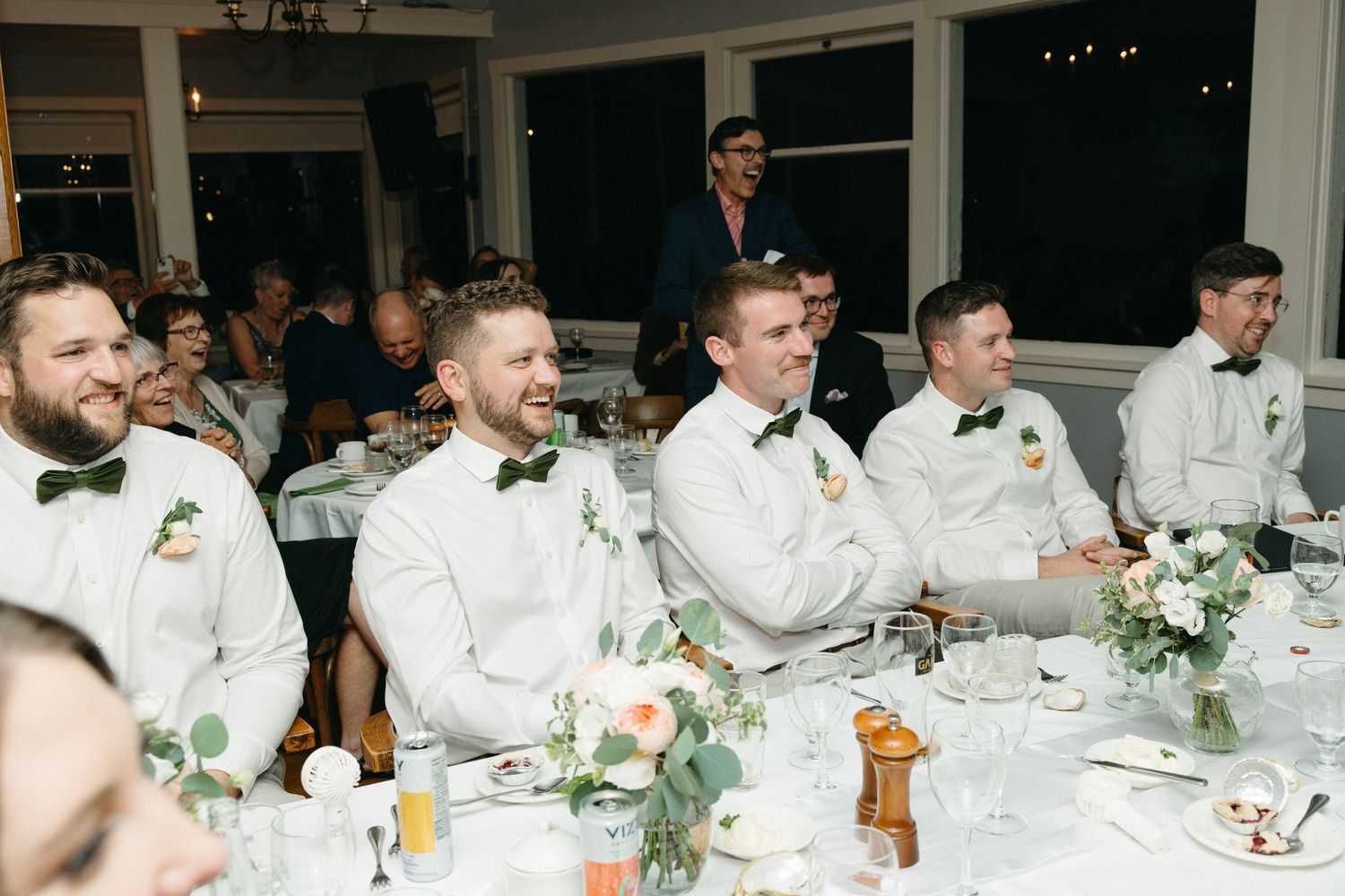 Groomsmen in white suits with green bow ties laugh and smile during wedding reception speeches at decorated table.