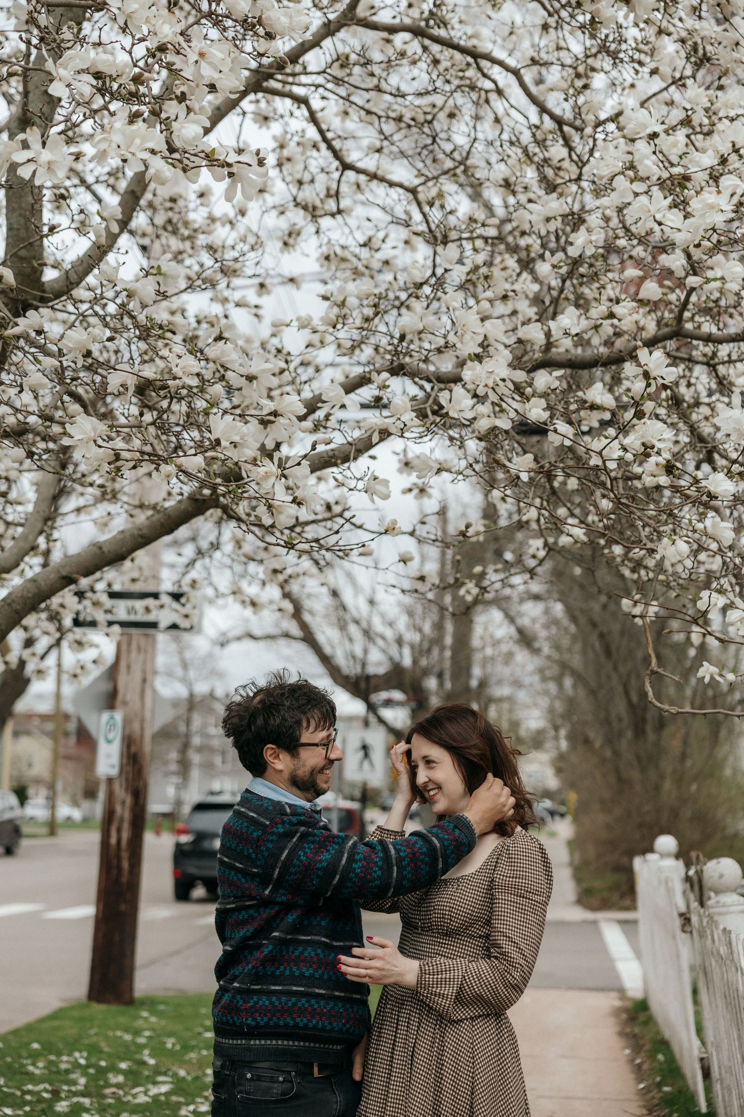 A couple embracing and smiling at each other under a blooming white magnolia tree on a sidewalk in spring.