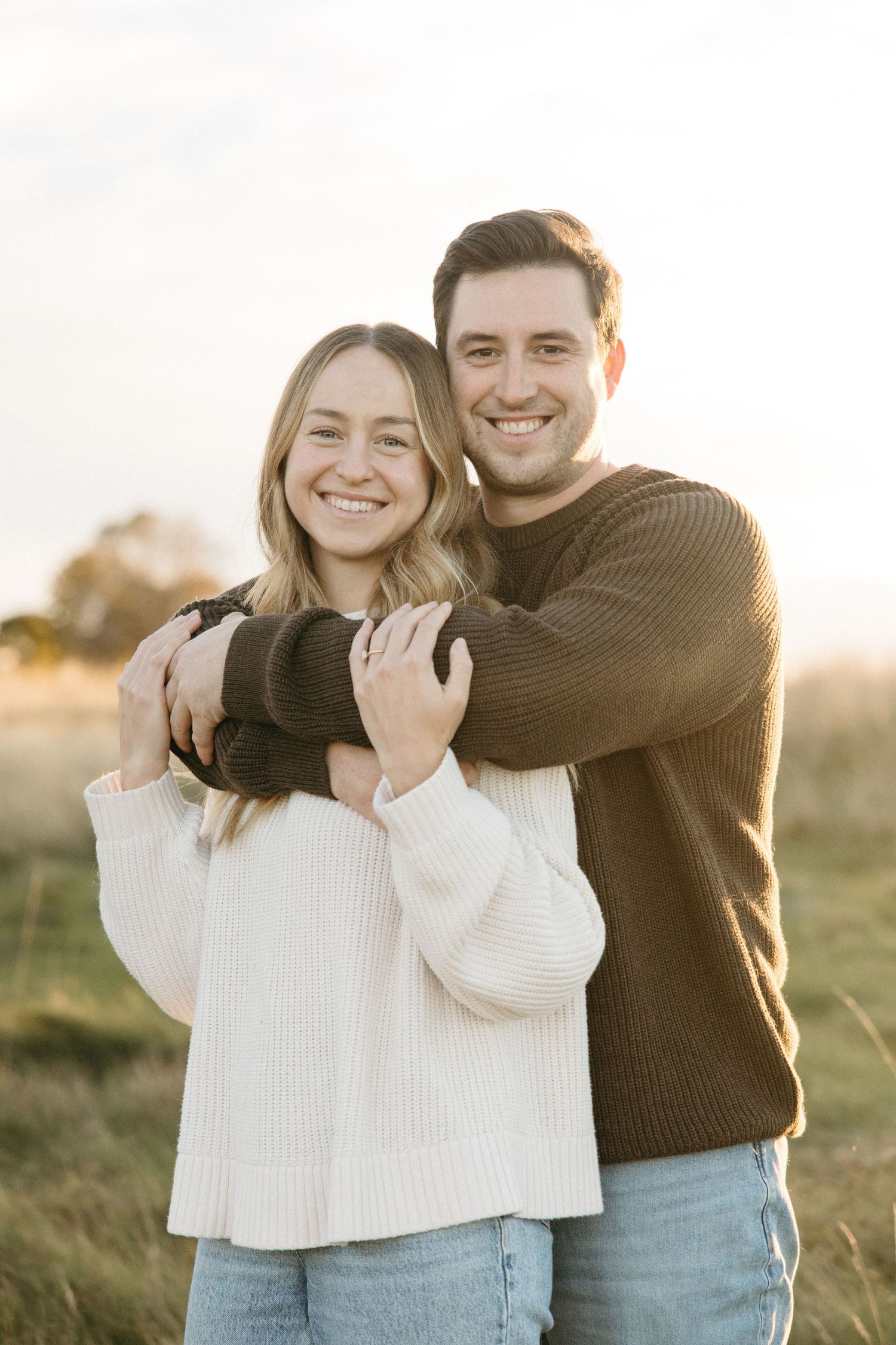 Smiling couple embracing outdoors in a grassy field at golden hour, she wears a cream knit sweater and he wears a brown sweater.