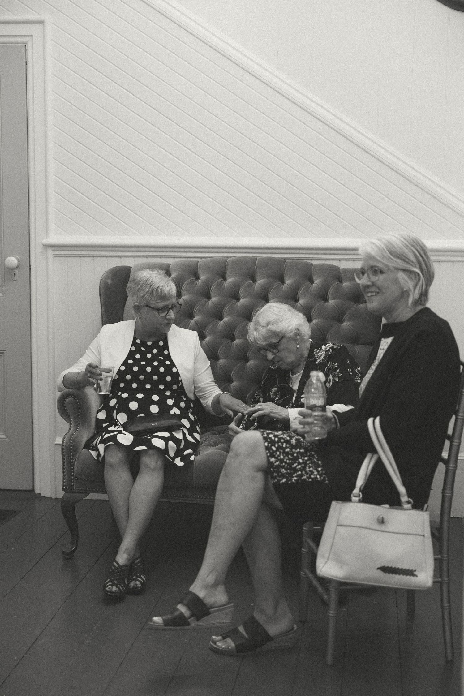 Three elderly women chat while sitting on a tufted gray couch at a formal event.
