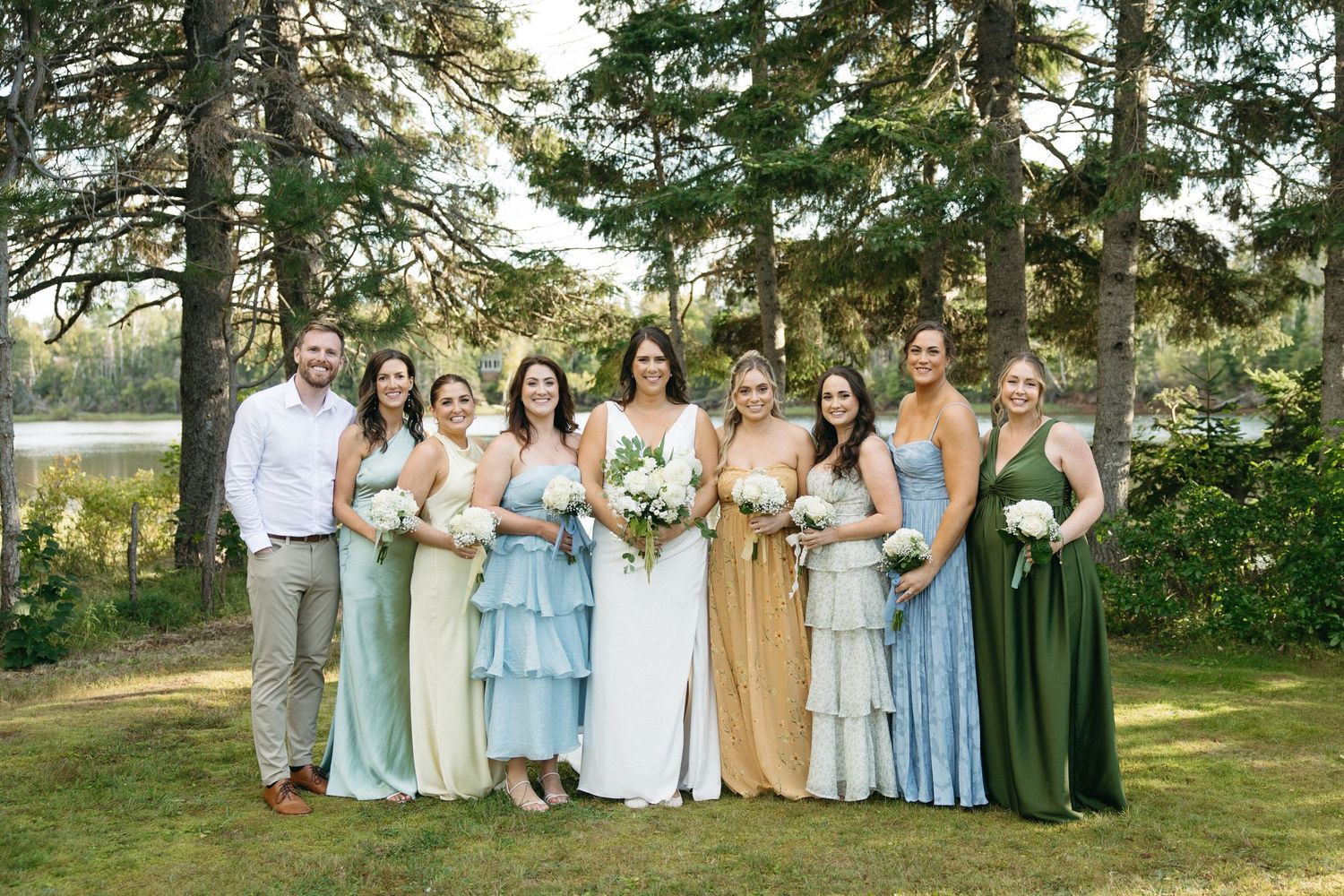 Large wedding party group photo with bridesmaids in coordinated pastel dresses outdoors.