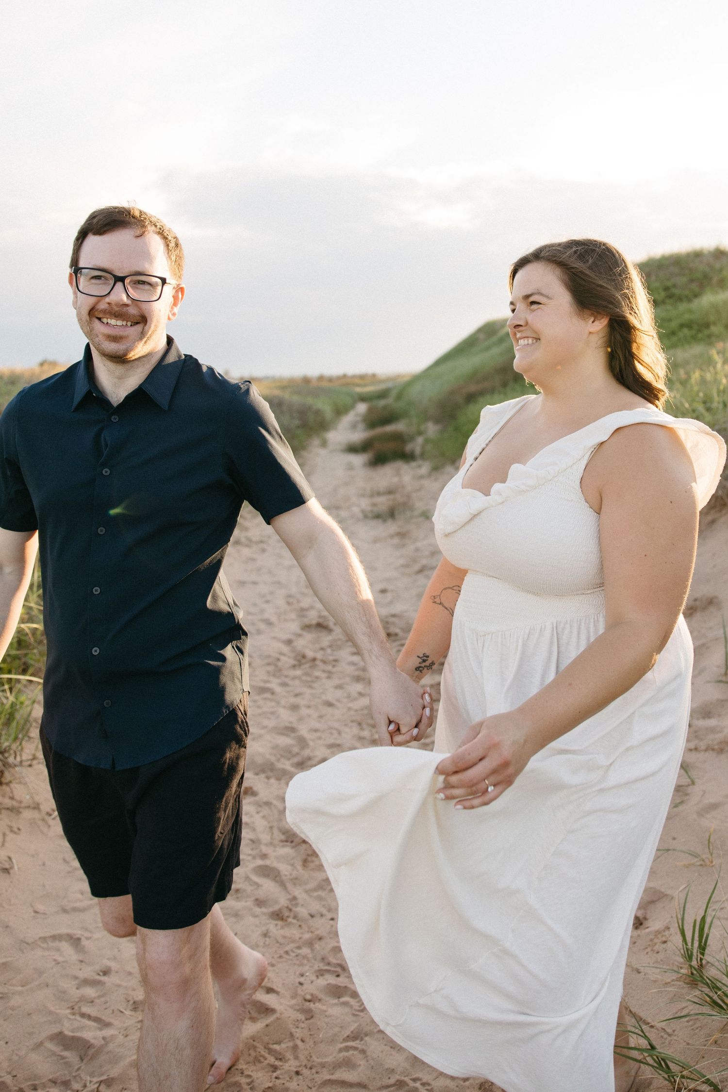 Happy couple holding hands walking on sandy beach path at sunset.