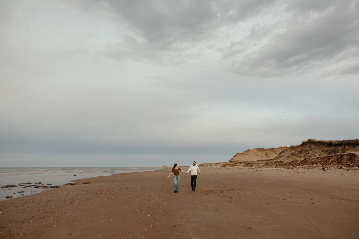 Two people walk along a misty beach shoreline under overcast skies with sand dunes in the distance.
