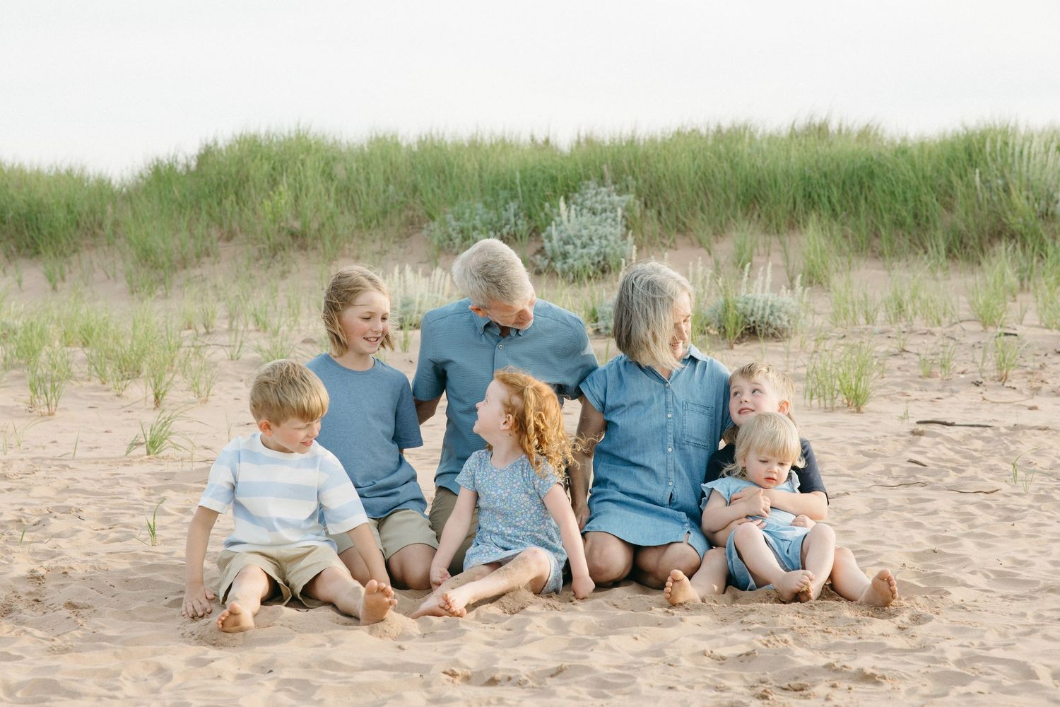 Grandparents sit on sandy beach surrounded by grandchildren in coordinating blue outfits.