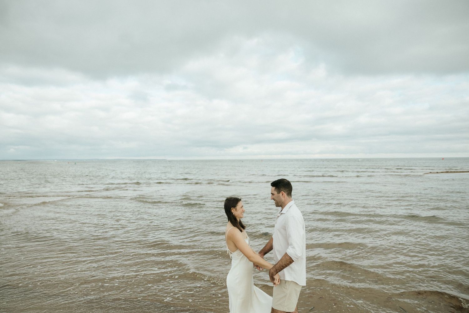Two people in white clothing dance playfully at the water's edge on a cloudy beach.