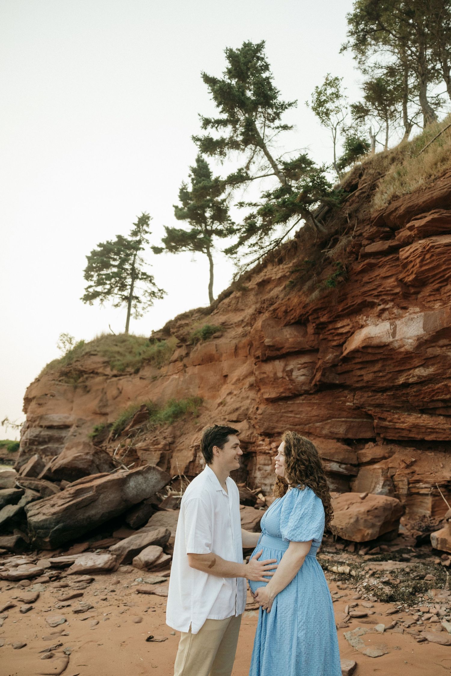 A couple in coordinating blue and white outfits pose against dramatic red cliffs at the beach.