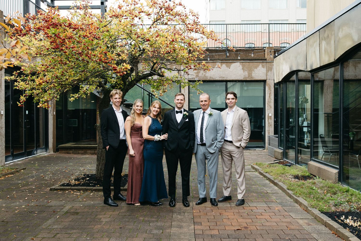A group in formal attire poses for a photo in a courtyard with fall foliage overhead.