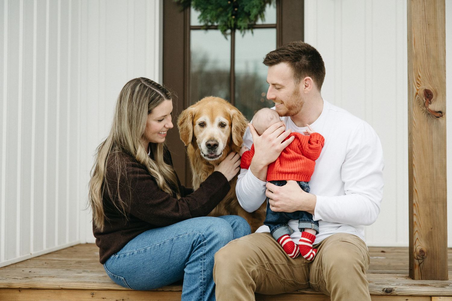 Cozy family portrait with golden retriever wearing festive red bow on front porch.