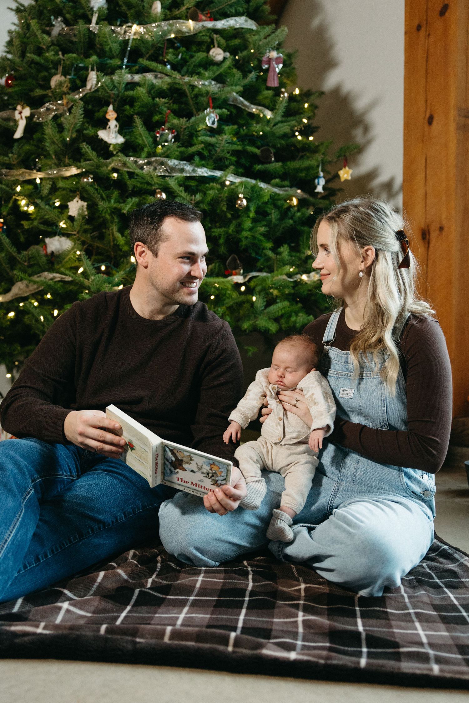 Family sharing intimate moments while reading on blanket beside decorated Christmas tree.