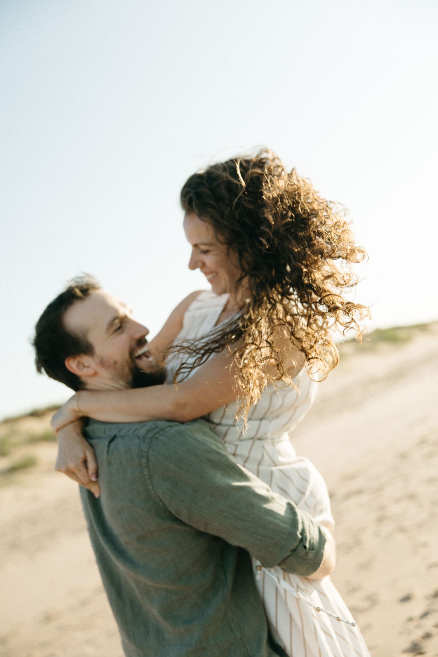 Joyful couple embracing on sunny beach, man lifting smiling woman with curly hair against blue sky.