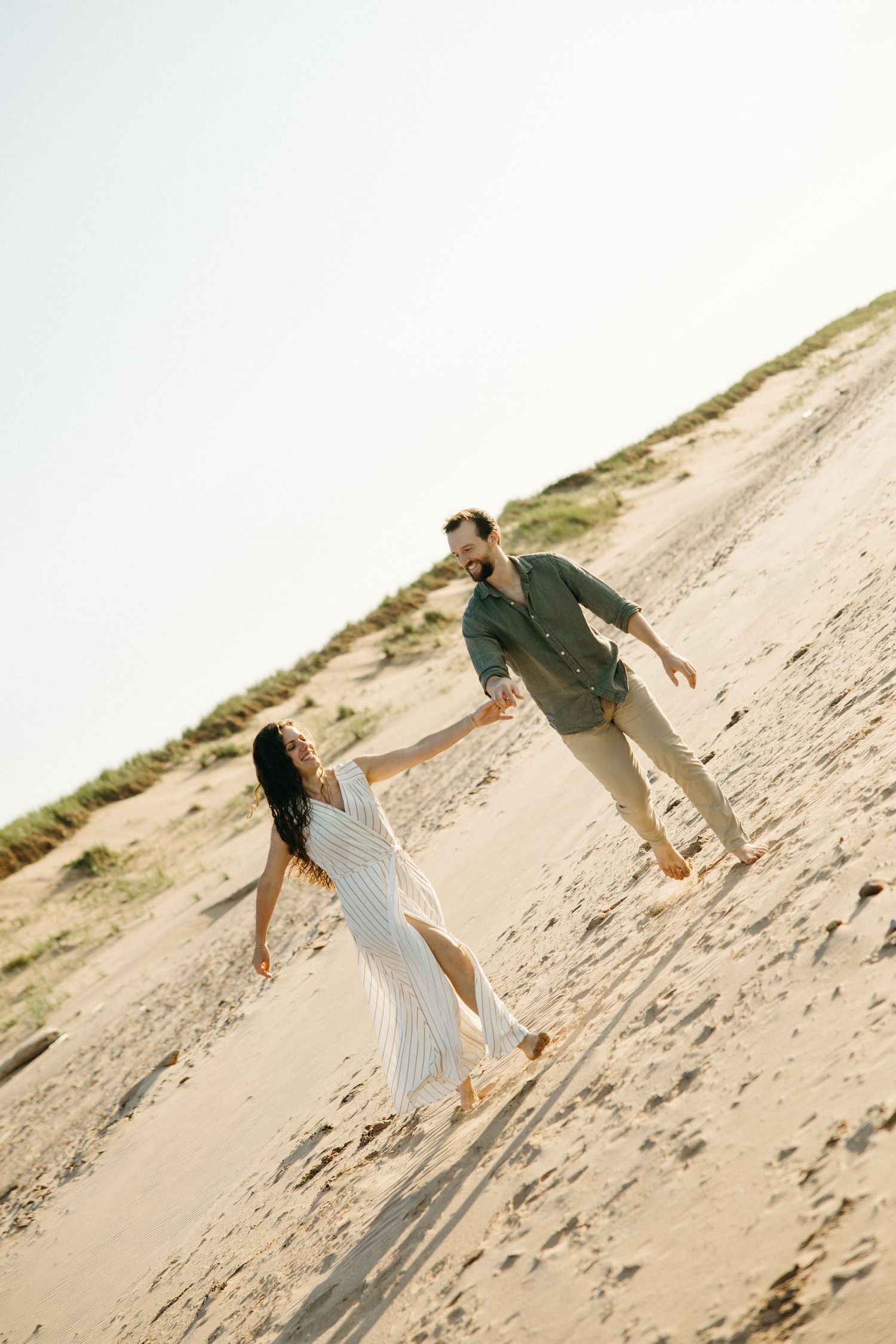 Couple holding hands and walking barefoot on sandy beach dunes in casual summer outfits.