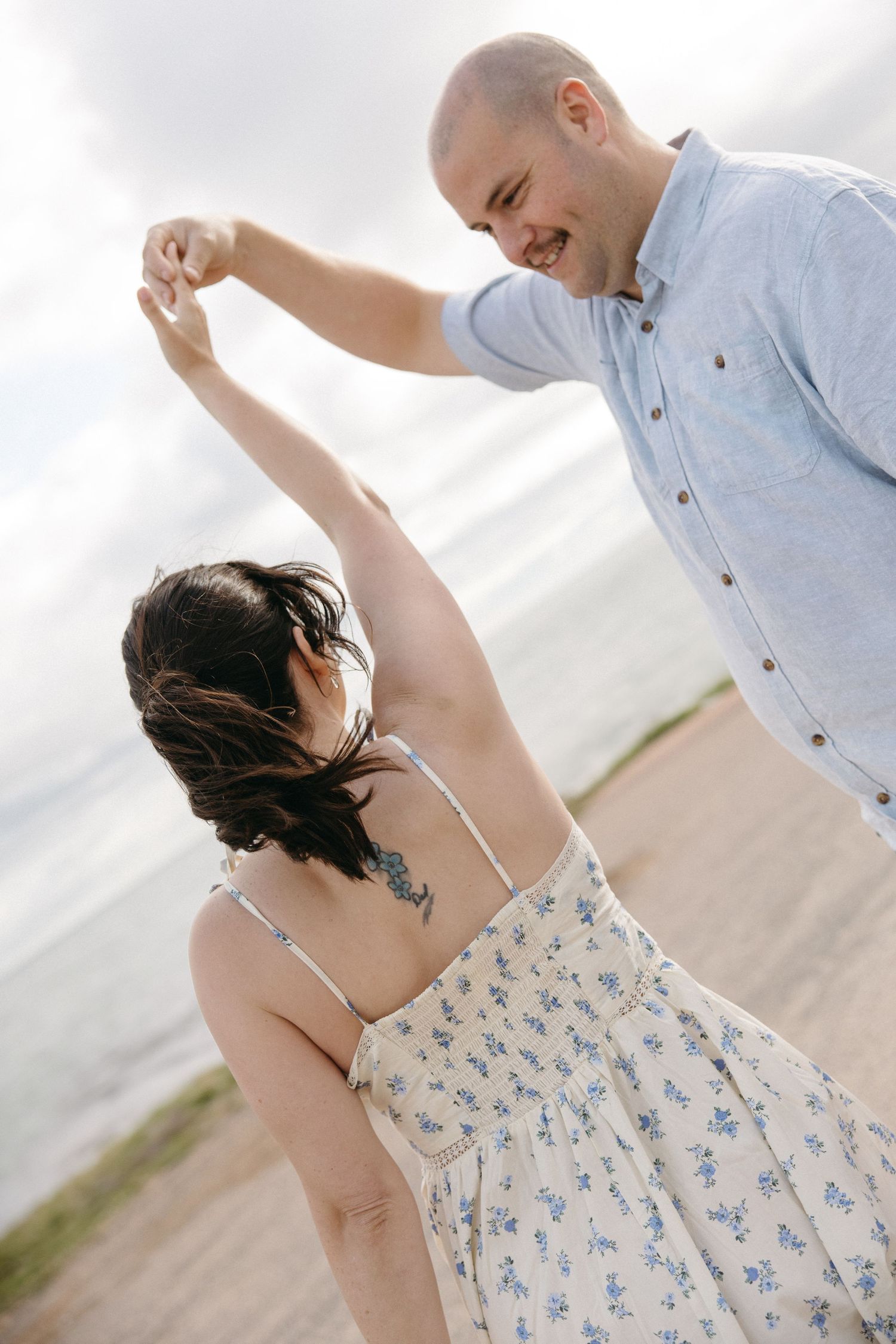 Smiling bald man spinning a dark-haired woman in a floral dress on a sandy beach.