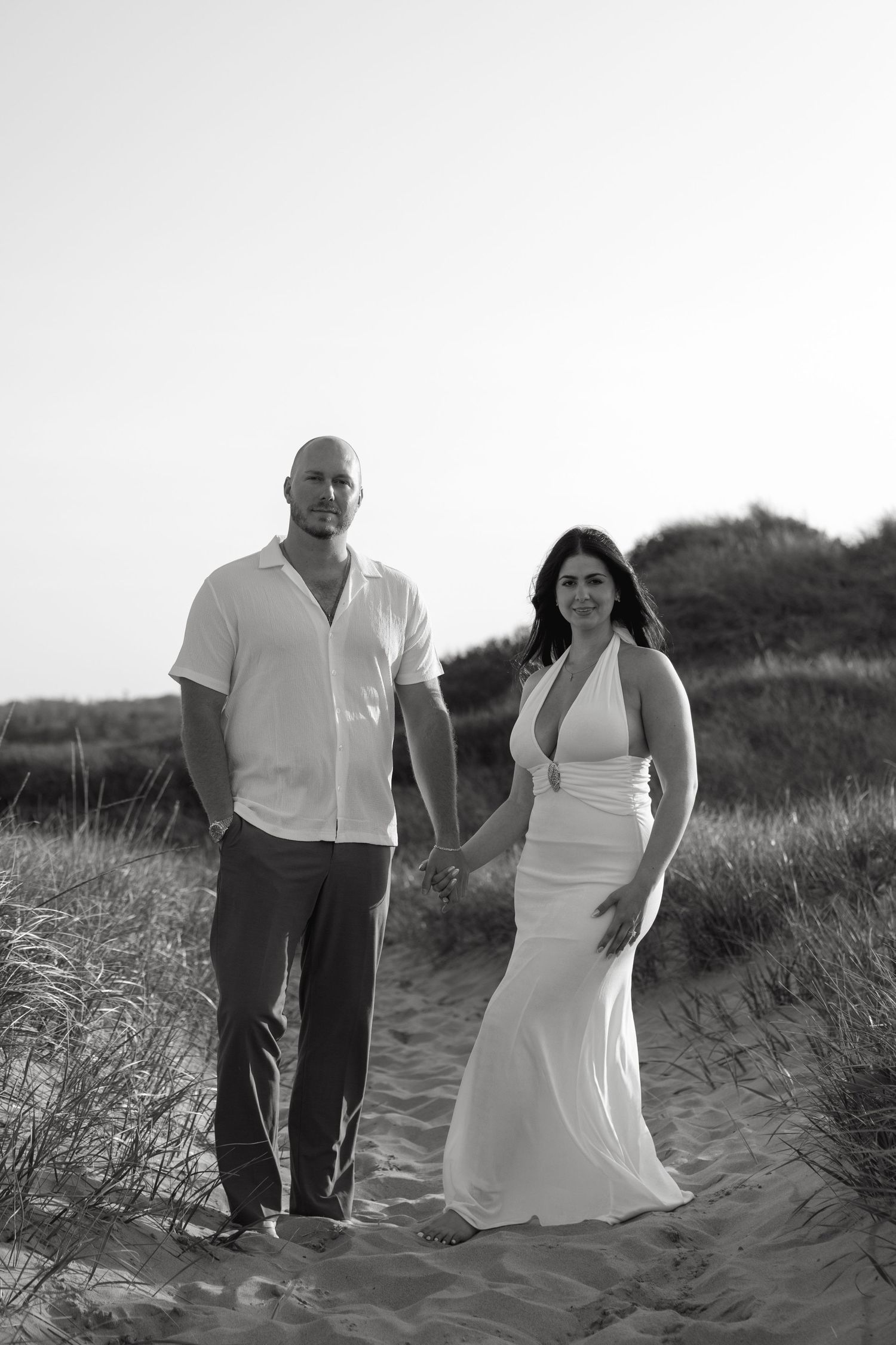 Black and white photo of couple holding hands on sandy beach path surrounded by grass dunes.