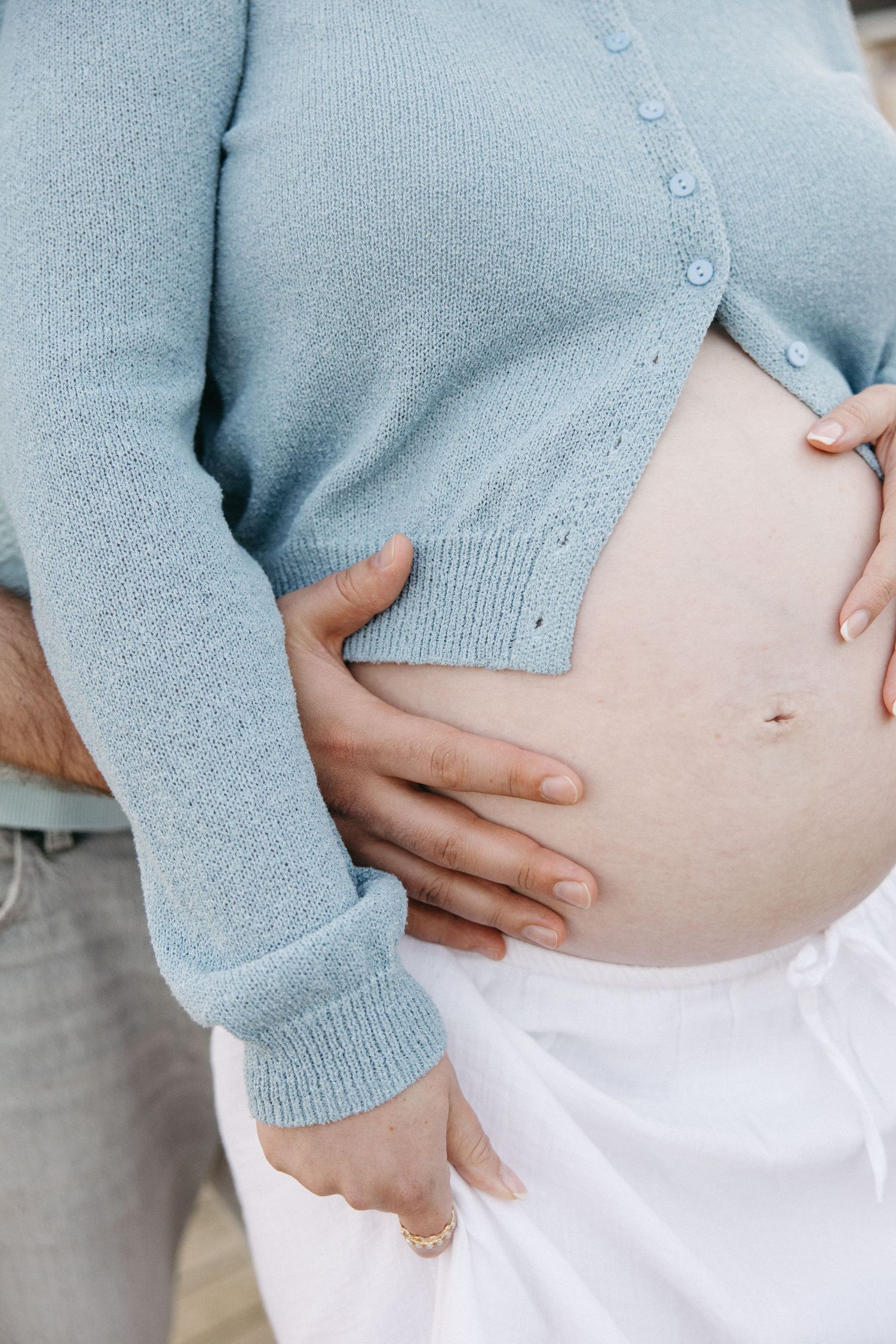 Close-up of pregnant woman in light blue cardigan cradling her bare baby bump with both hands.