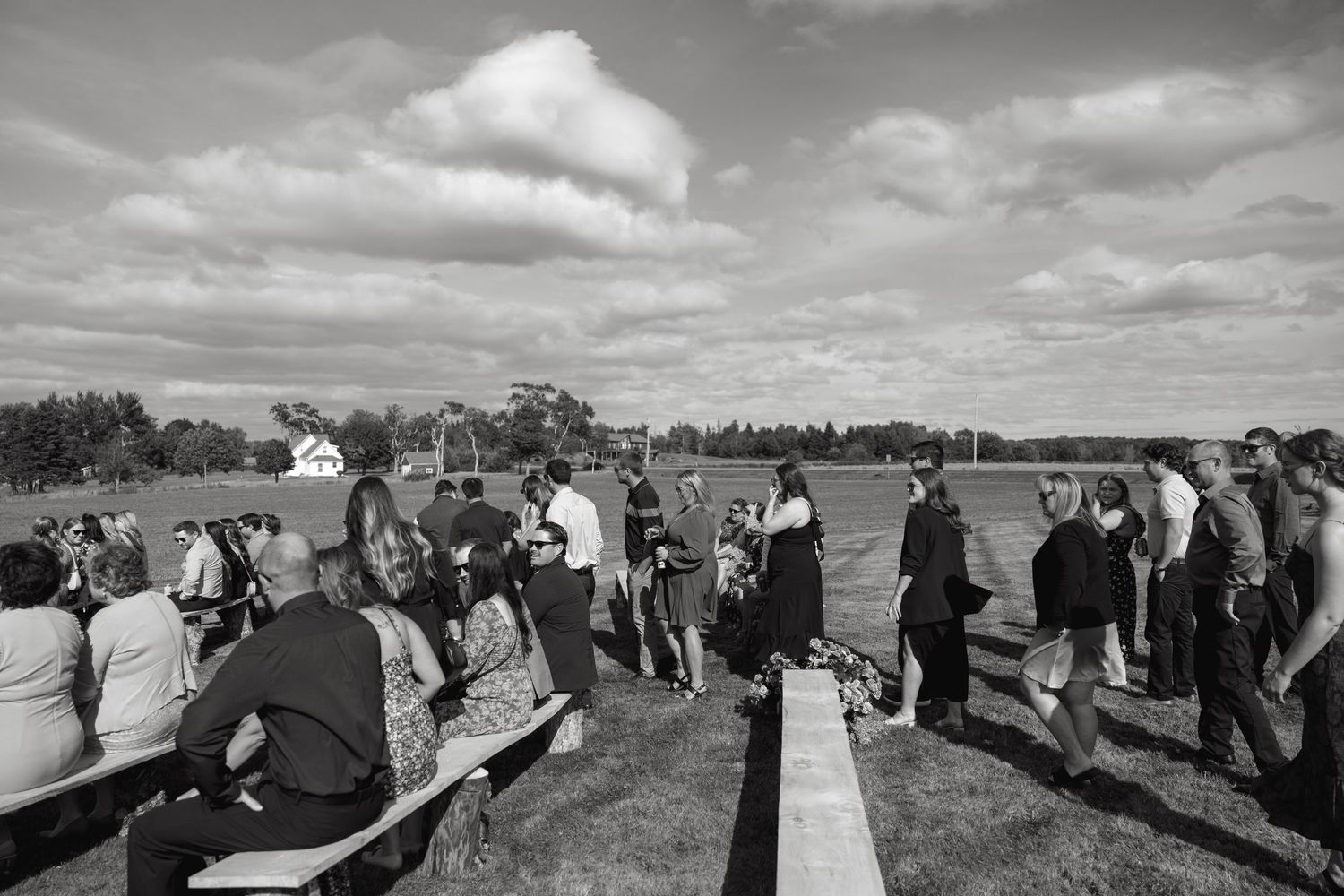 Wedding ceremony guests gather on rolling hills under cloudy sky.