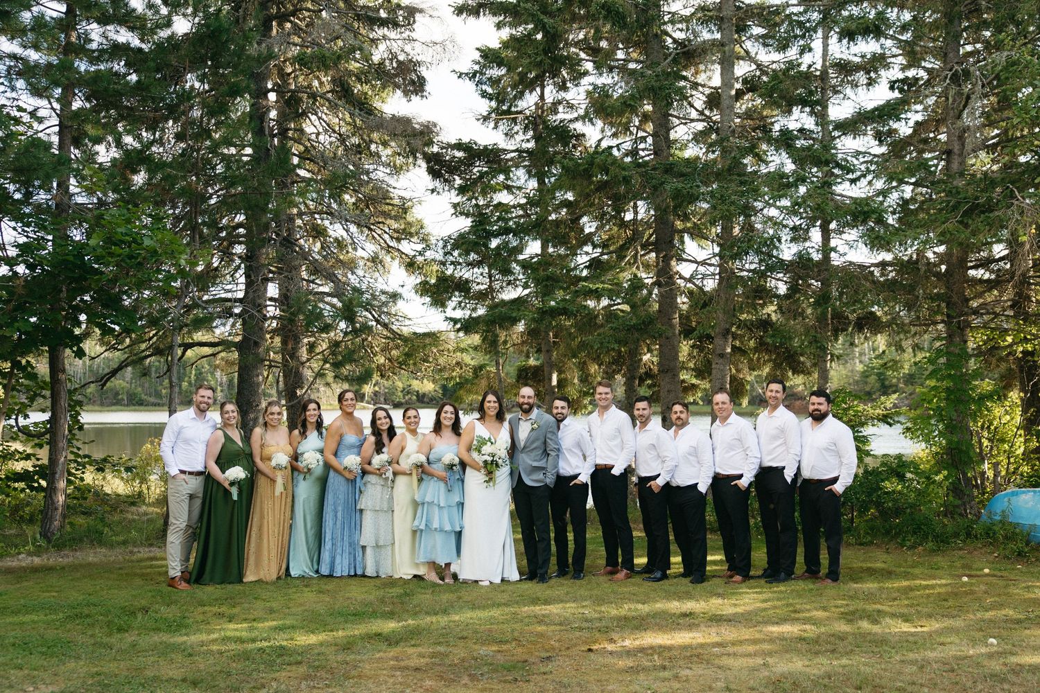 Wedding group gathers for formal photos on grassy hillside with evergreen trees in background.