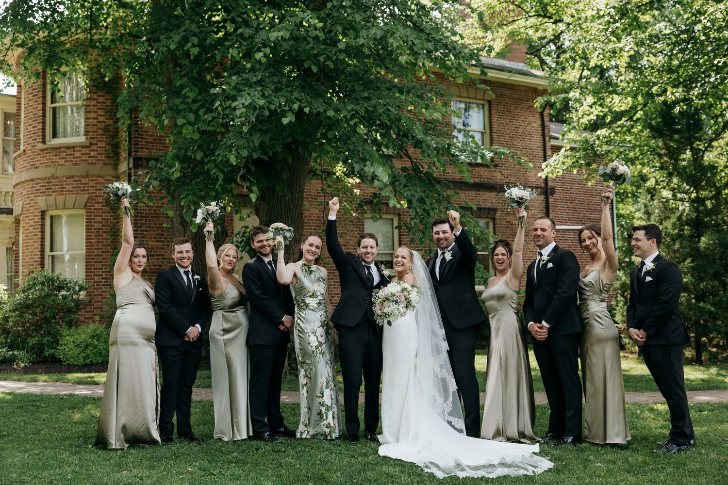 Full wedding party poses for formal group photos on green lawn near historic brick building.