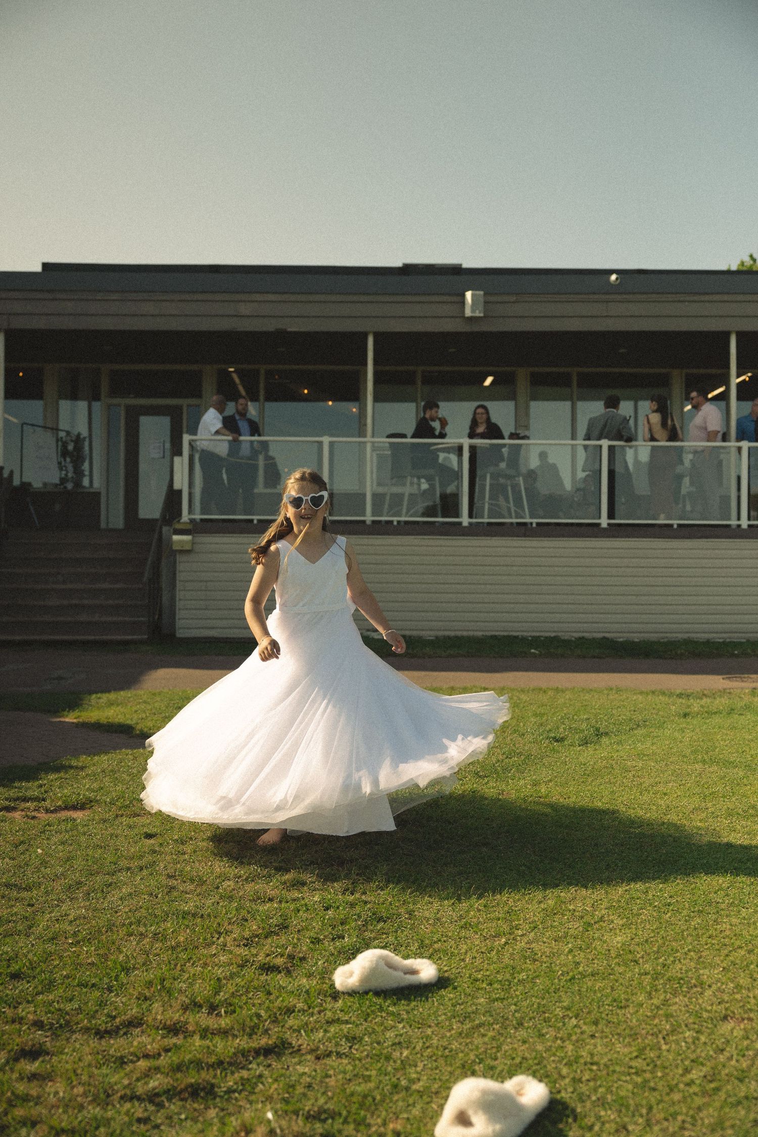Person in a flowing white wedding dress twirling on a grassy lawn near a modern building.