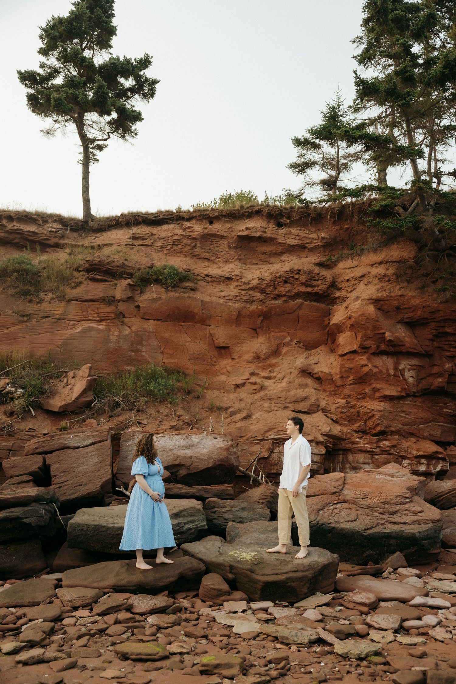 Two people pose for photos on rocky terrain with red cliffs and pine trees in the background.