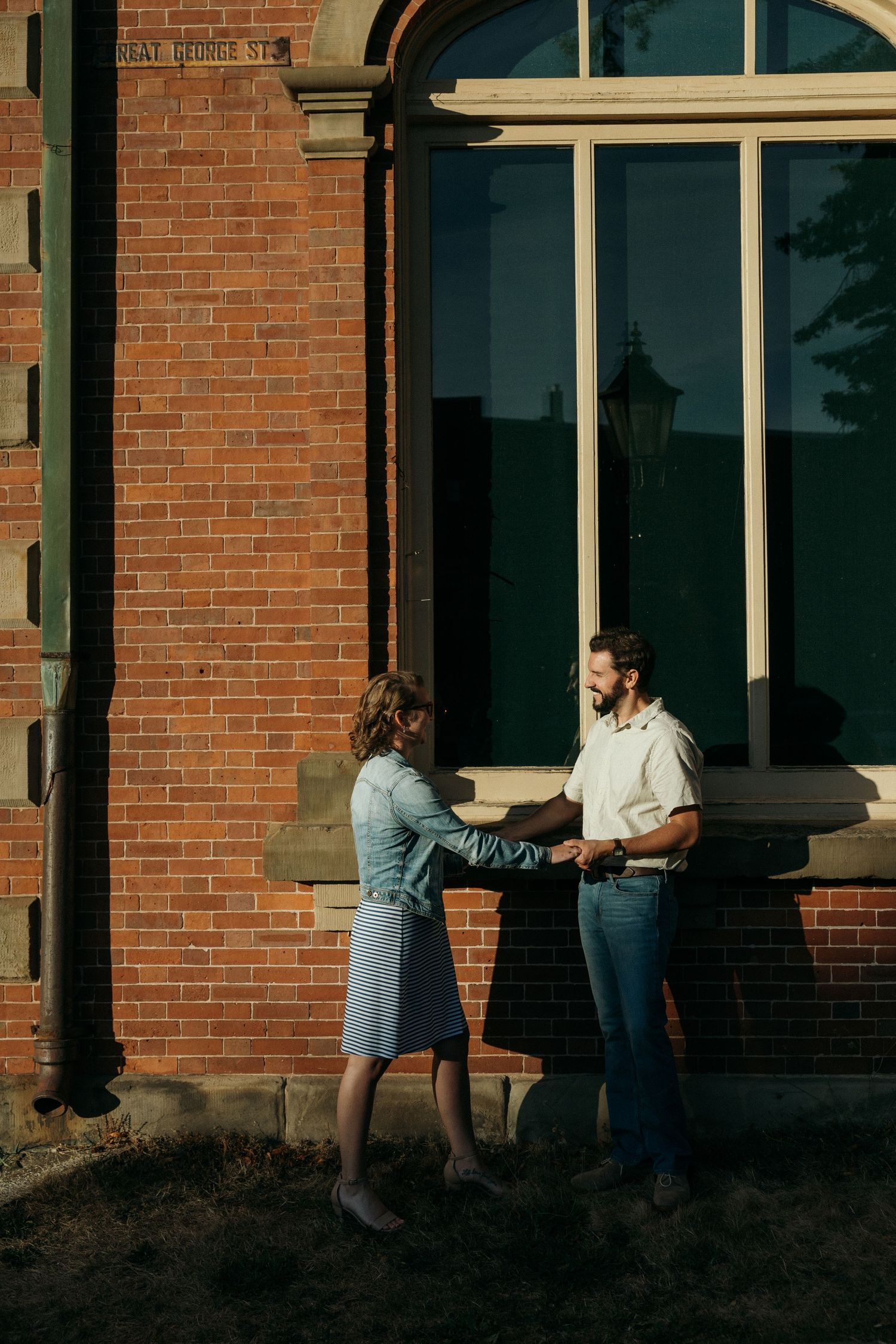 Two figures interact in golden sunlight against a brick building facade.