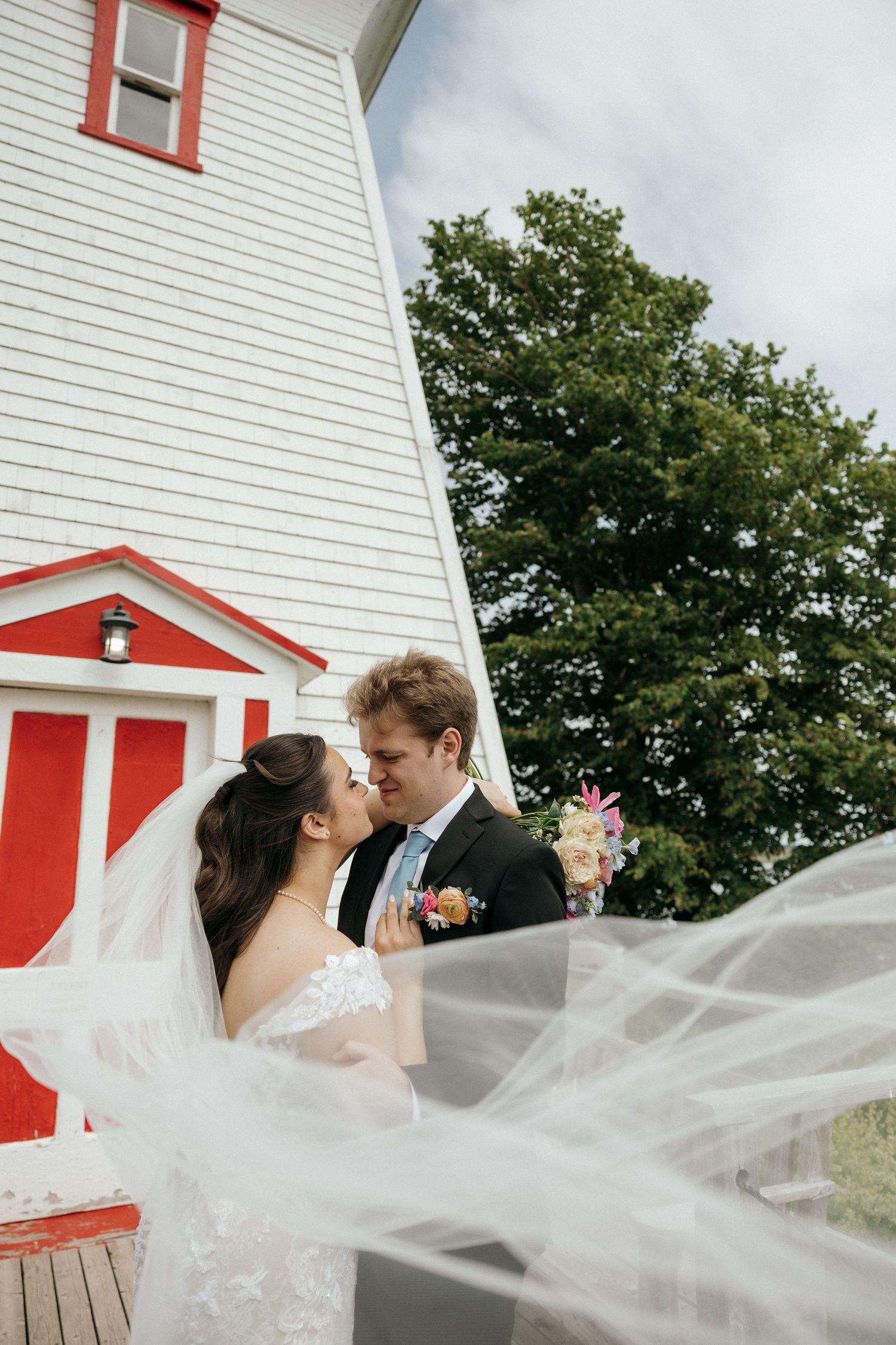 A flowing veil catches the breeze in front of a white and red lighthouse.