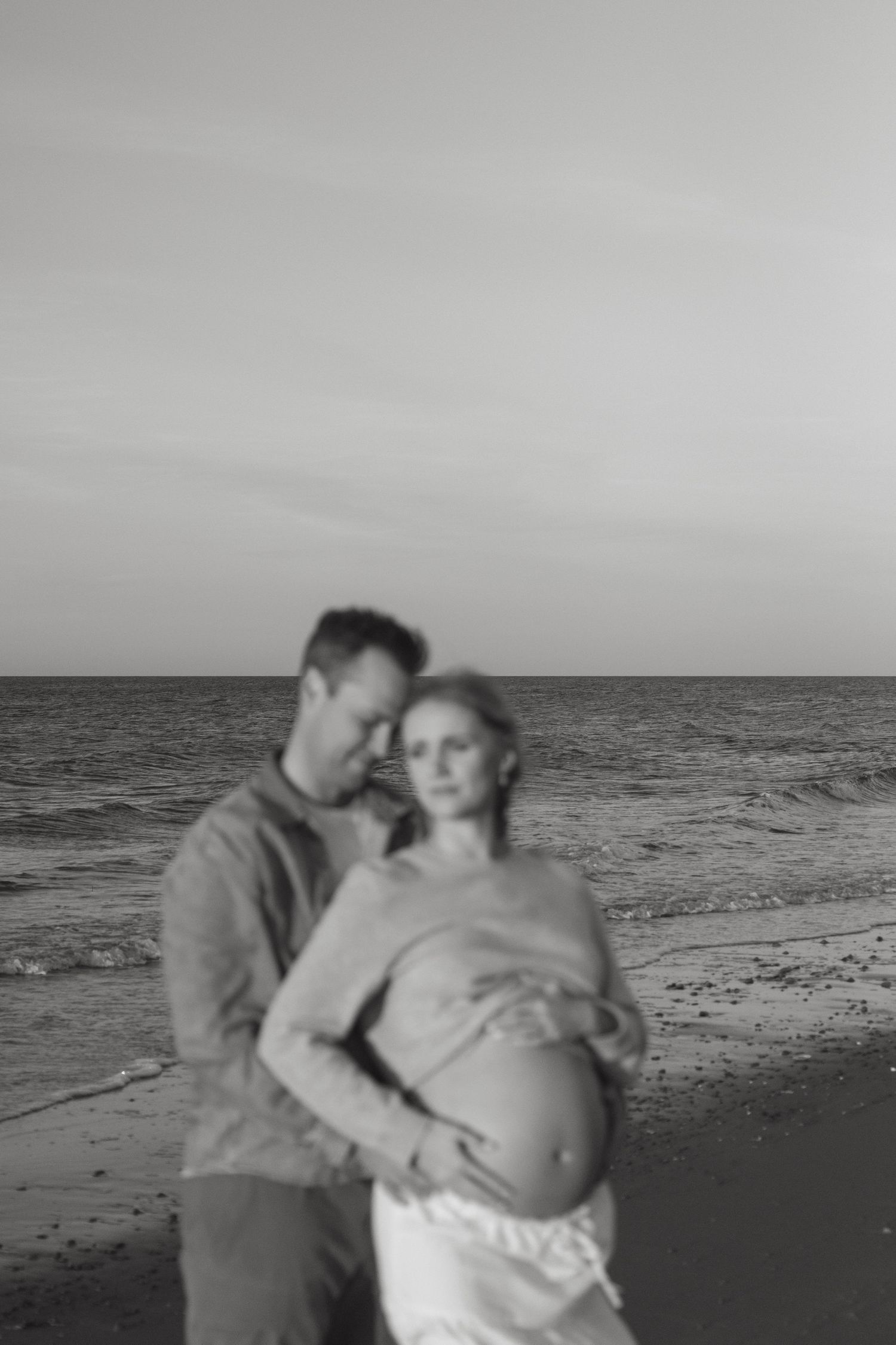 A black and white beach portrait of an expecting couple embracing by the ocean.