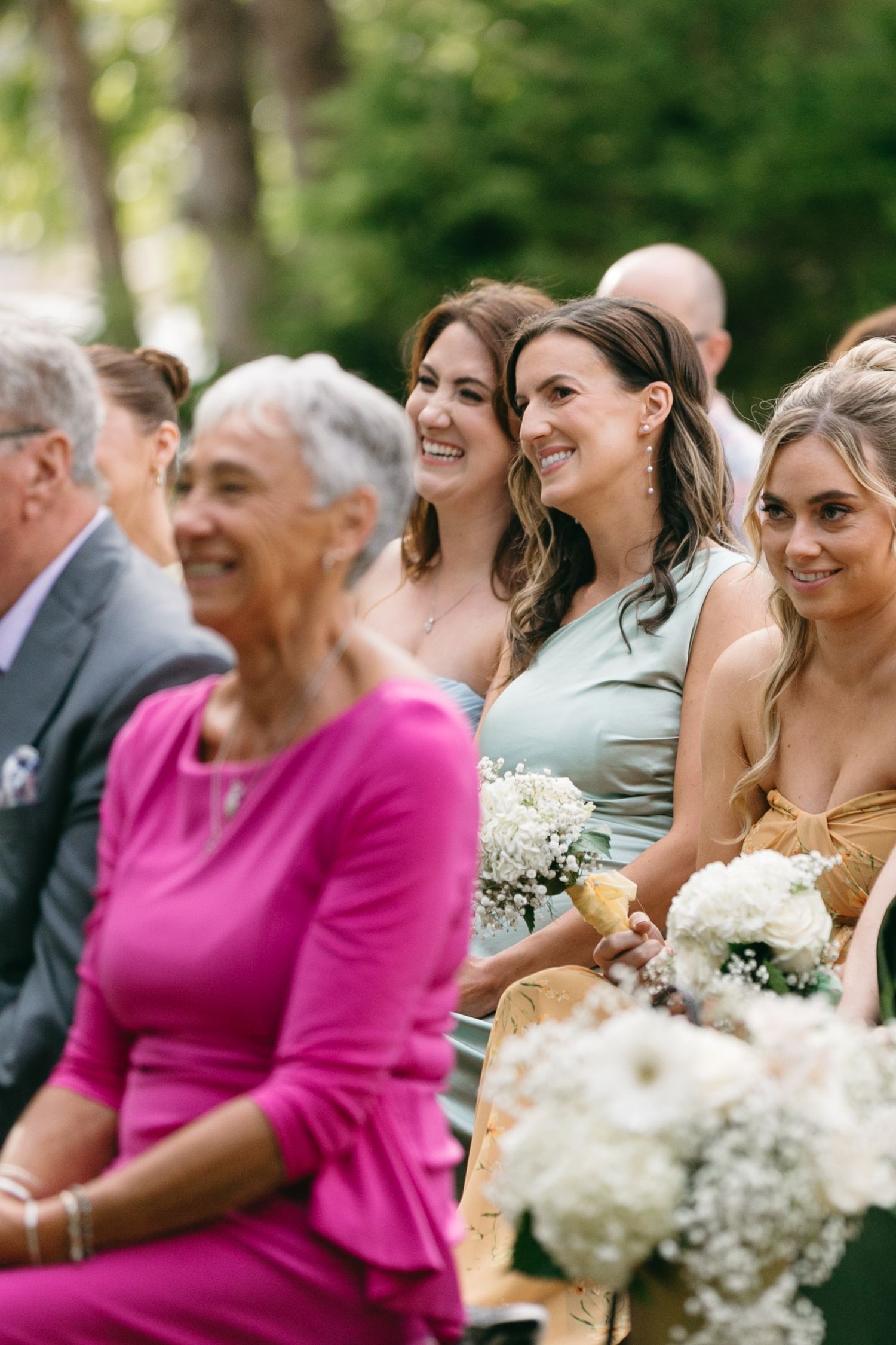 Wedding party members and guests smile during an outdoor ceremony with floral bouquets.