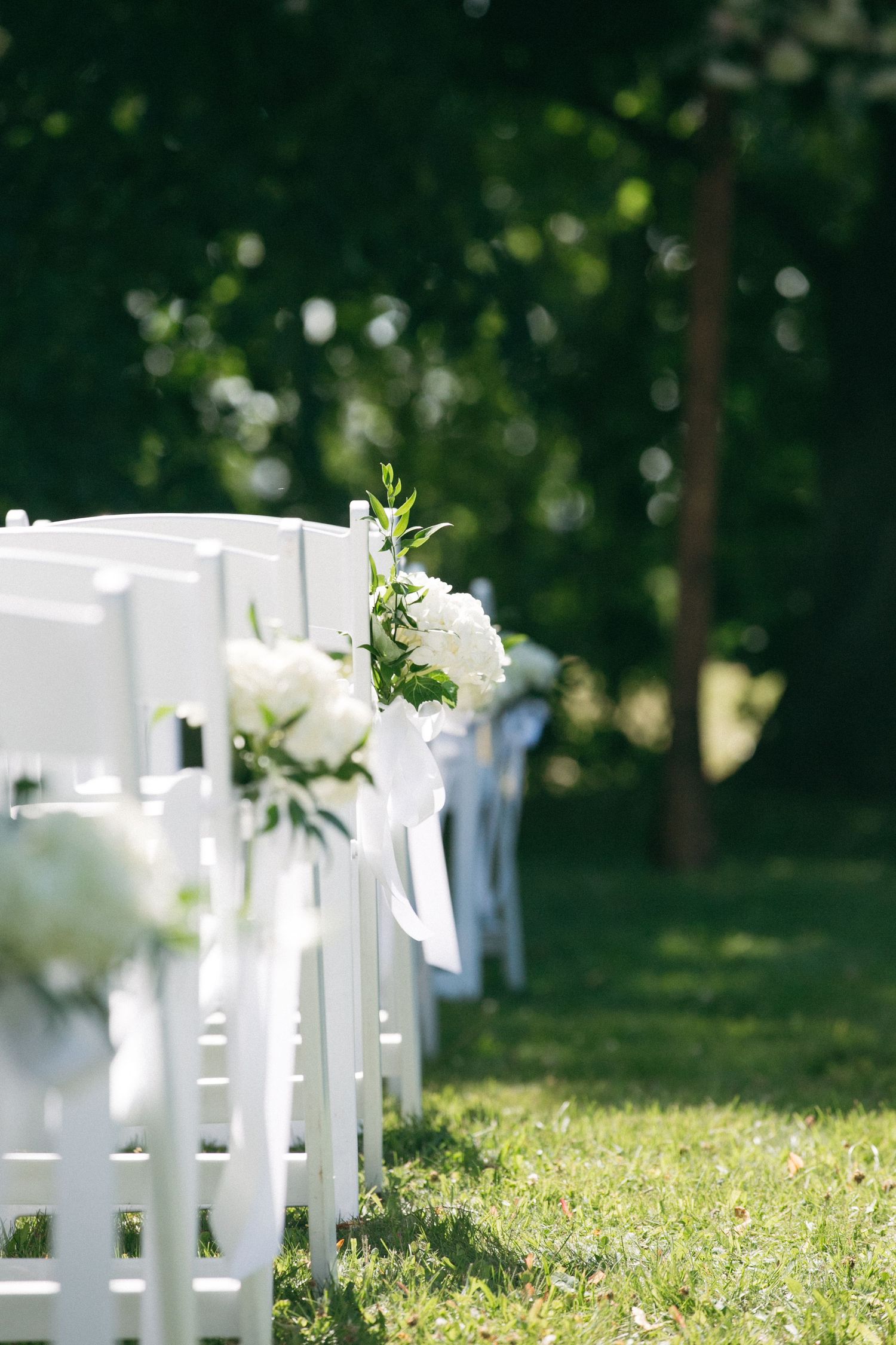 White ceremony chairs decorated with greenery arrangements in an outdoor garden setting.