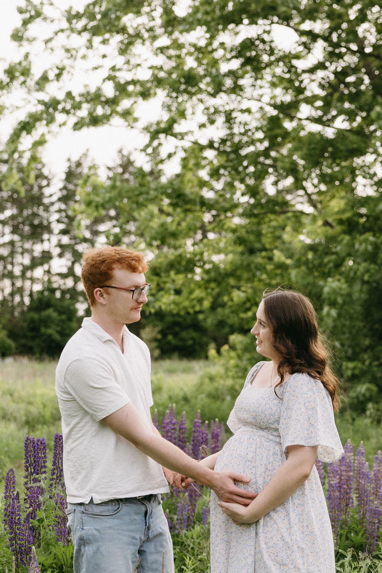 A couple in white outfits embrace and laugh together in a garden with blooming purple lupine flowers and greenery.