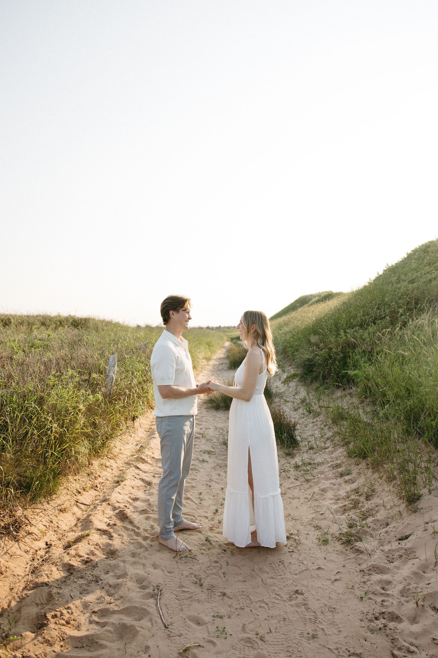 Couple holding hands facing each other on a sandy beach path surrounded by green dunes in golden light.