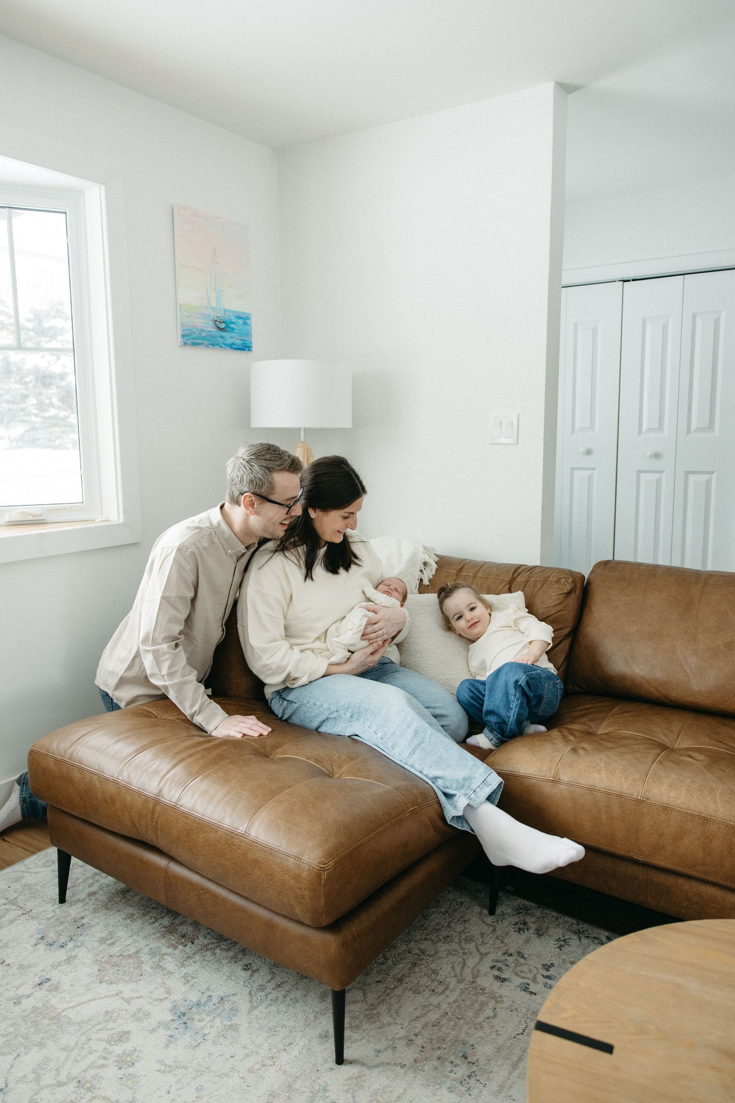 A family relaxes together on a brown leather sectional sofa in a bright, minimalist living room.