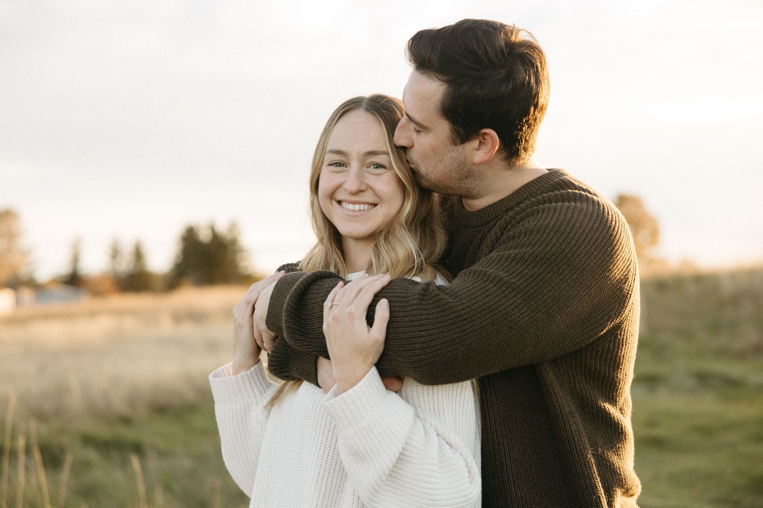 Smiling woman in a white knit sweater is embraced from behind by a man kissing her head in a grassy field at sunset.