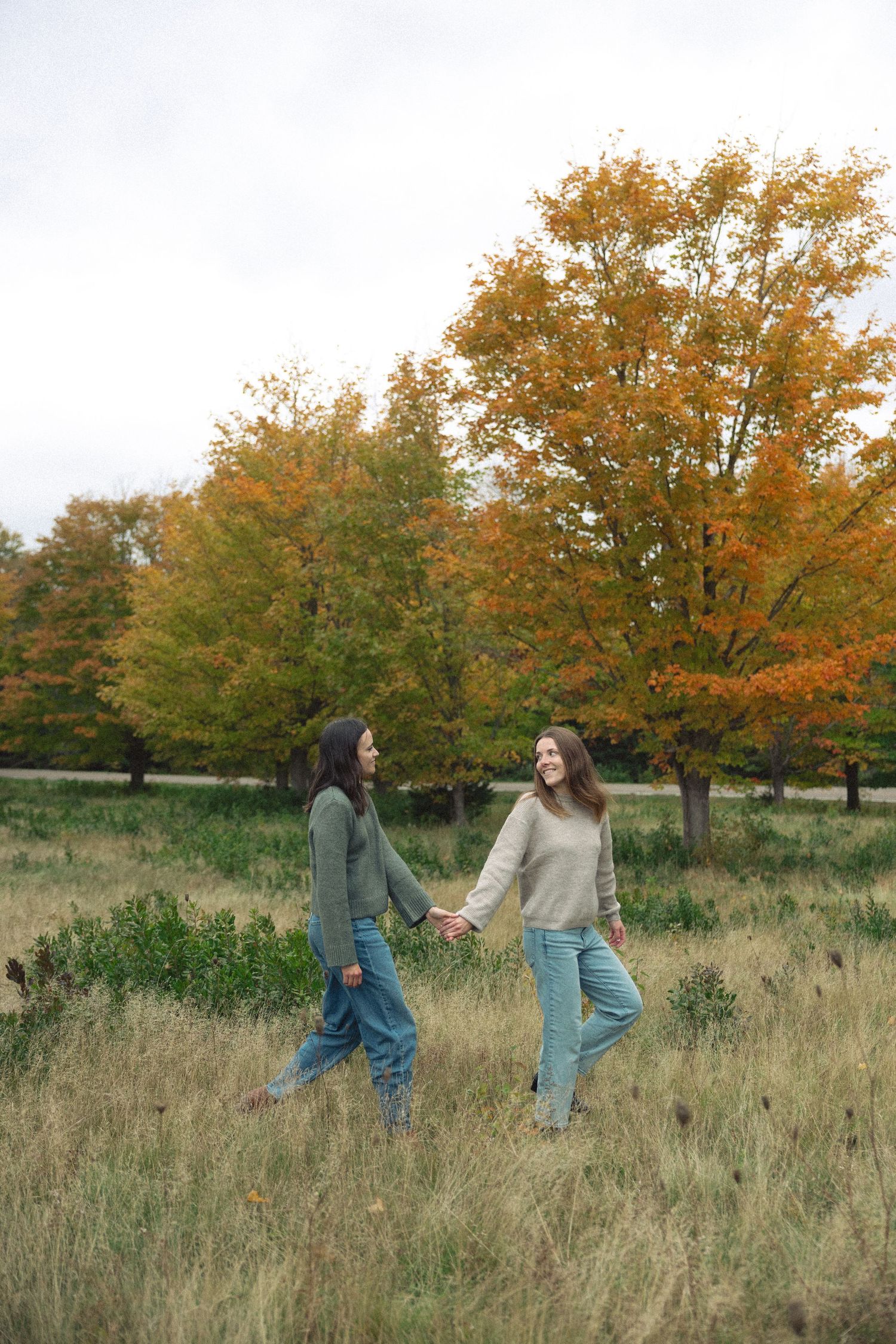 Two women hold hands and walk through a grassy field with colorful orange and yellow fall foliage in the background.