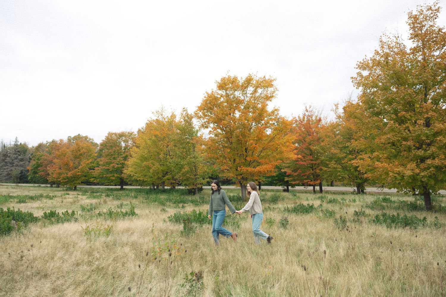 Two people hold hands and walk through a tall grass meadow beneath vibrant orange and yellow autumn trees on an overcast day.