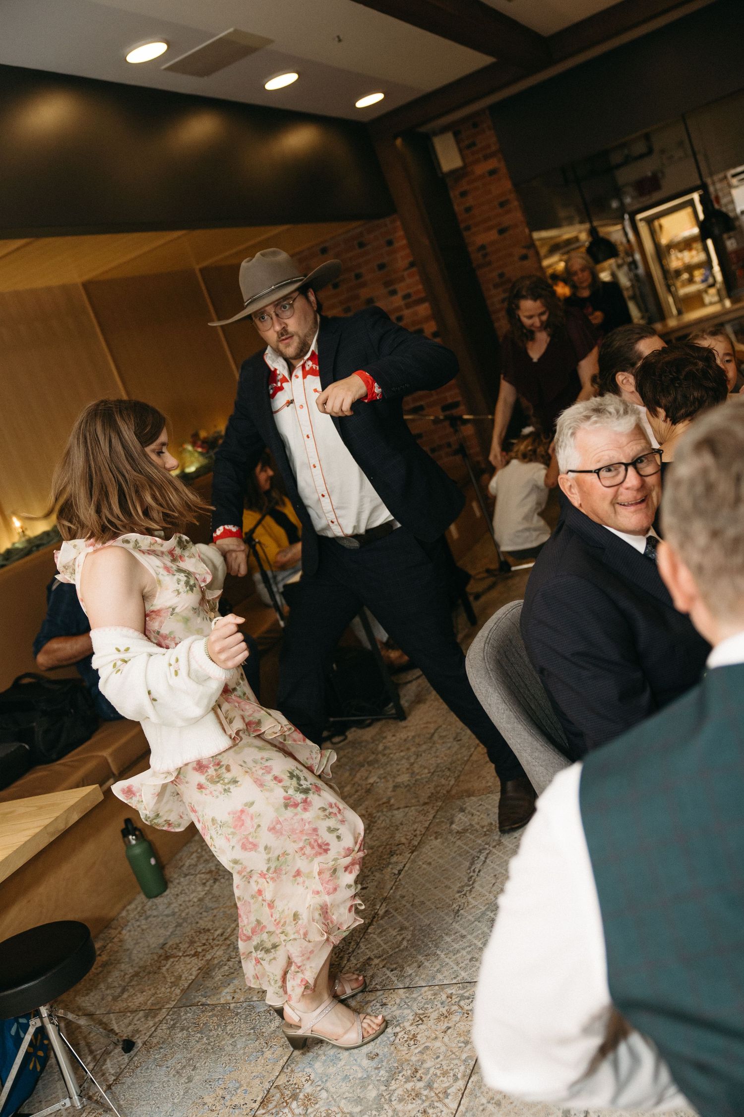 A man in a cowboy hat and bow tie dances with a woman in a floral dress while smiling guests watch at a lively indoor celebration.